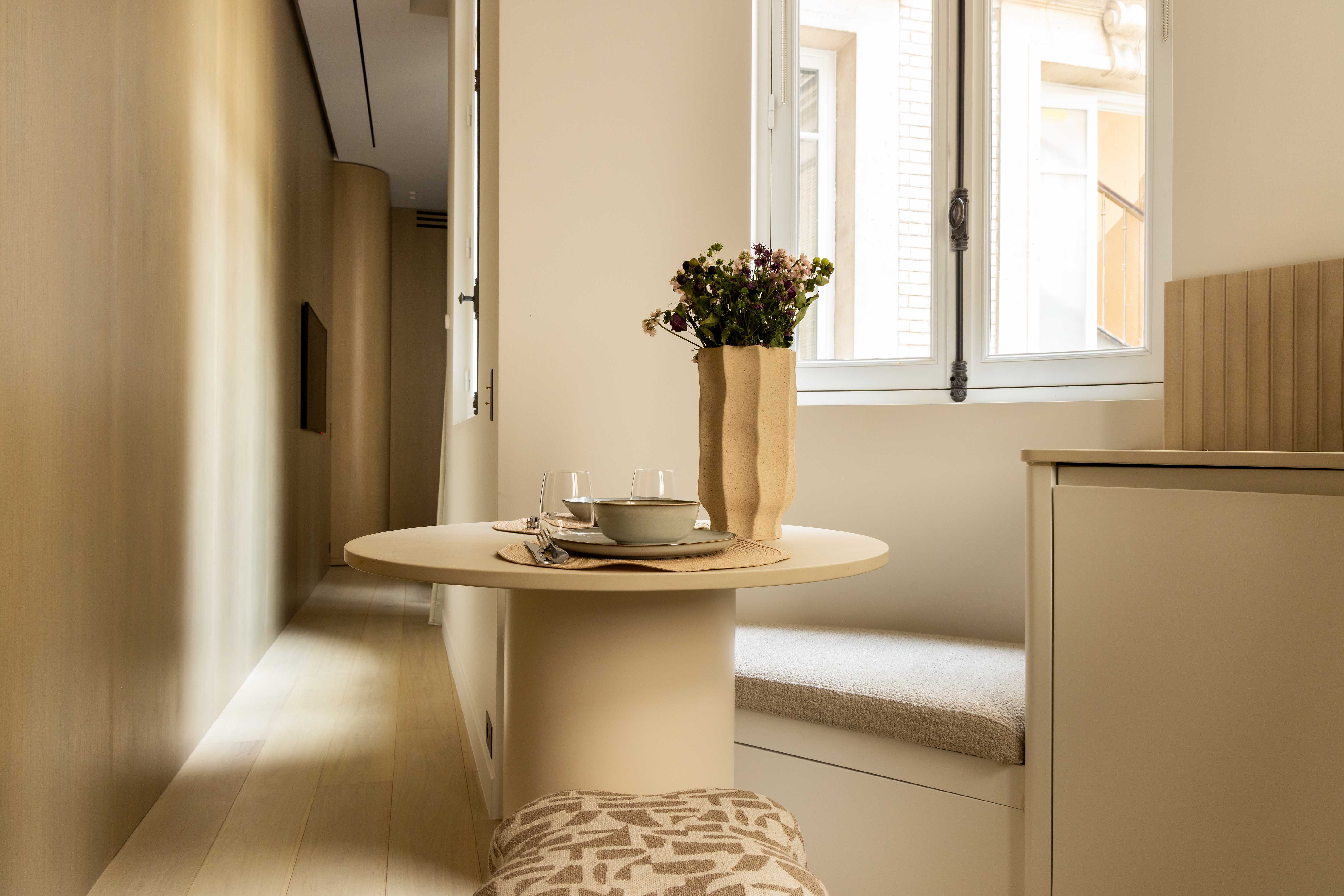 Coin repas avec petite table ronde beige, vaisselle, vase de fleurs, banquette et vue sur kitchenette