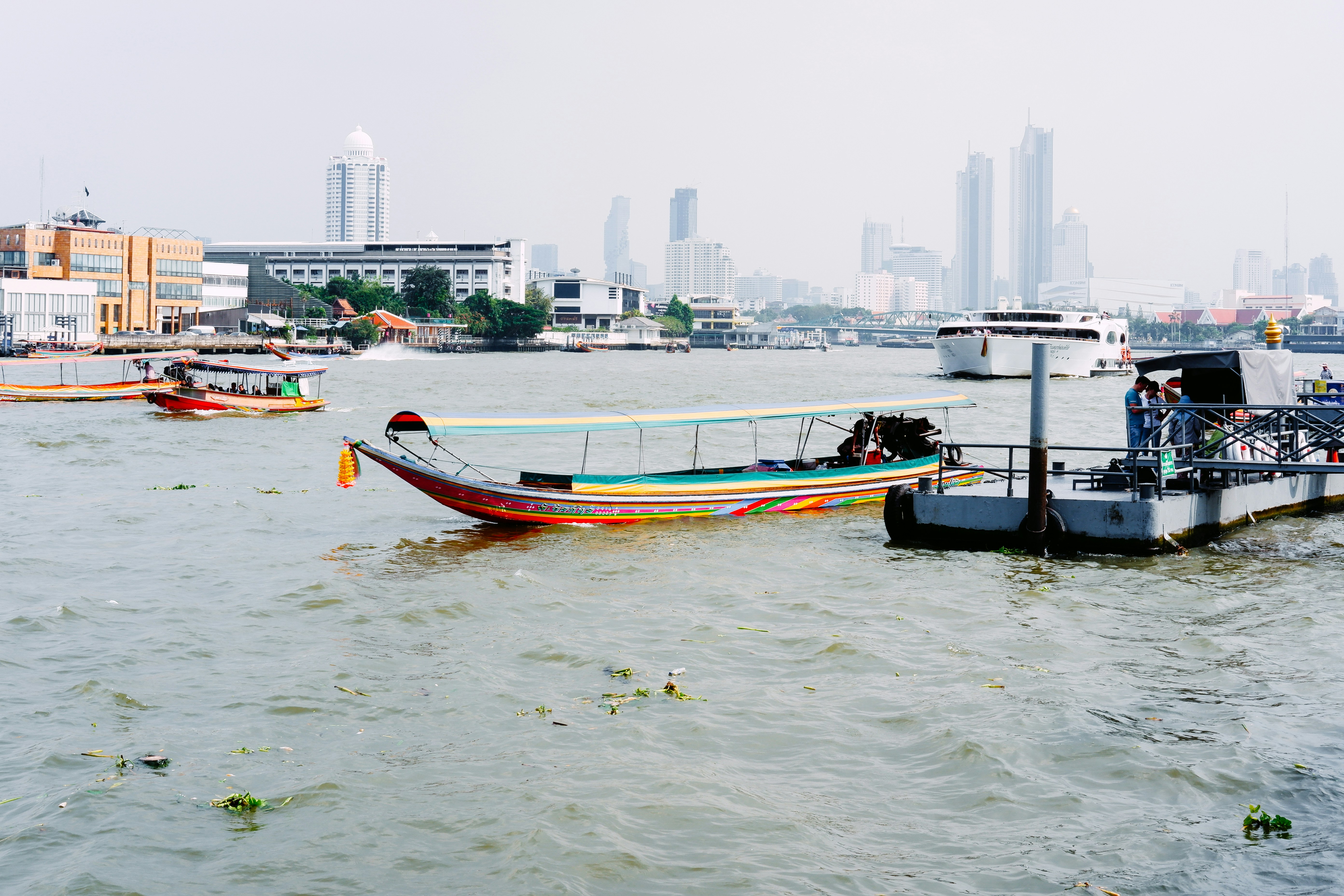 A colorful long-tail boat floats beside a small pier on a busy river, with several other boats in the water and modern city buildings visible through the haze in the background.