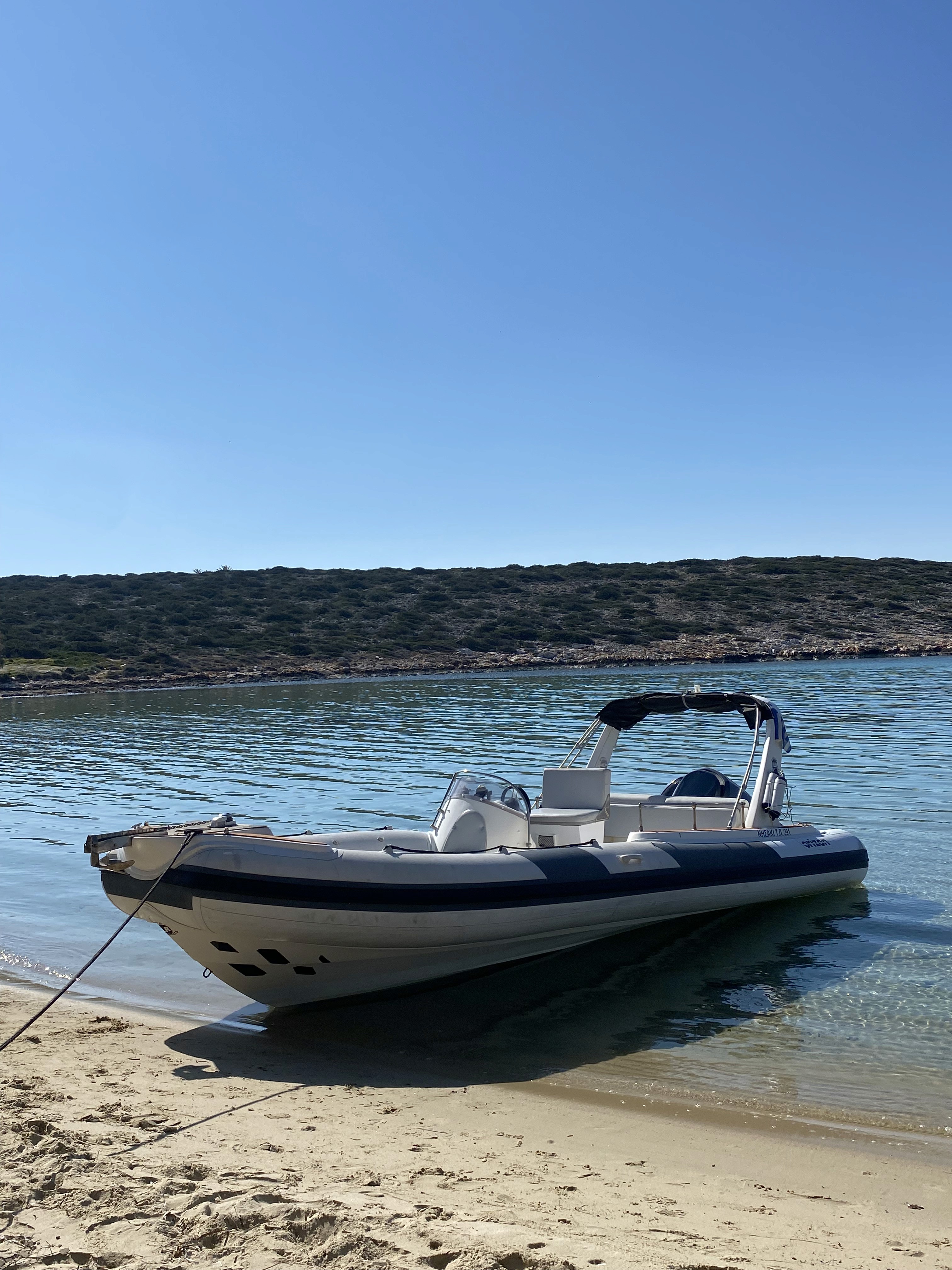 White and navy inflatable boat with center console beached on sandy shore in calm turquoise waters of a secluded Greek cove.