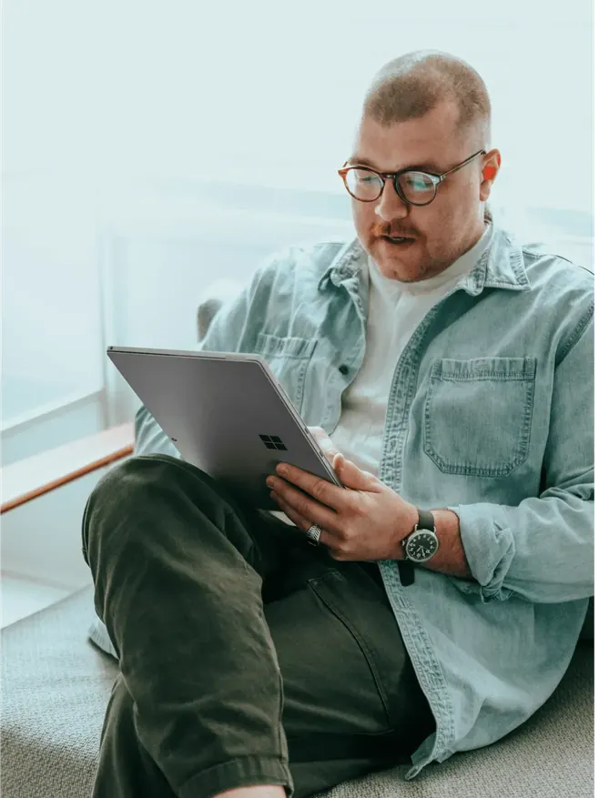Man with glasses using a tablet while seated on a sofa.