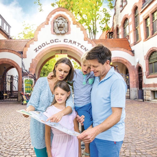 4人家族が石畳の道の上で、動物園の入り口前にある地図を見ています。そのアーチ形の門には「動物園」と記されています。