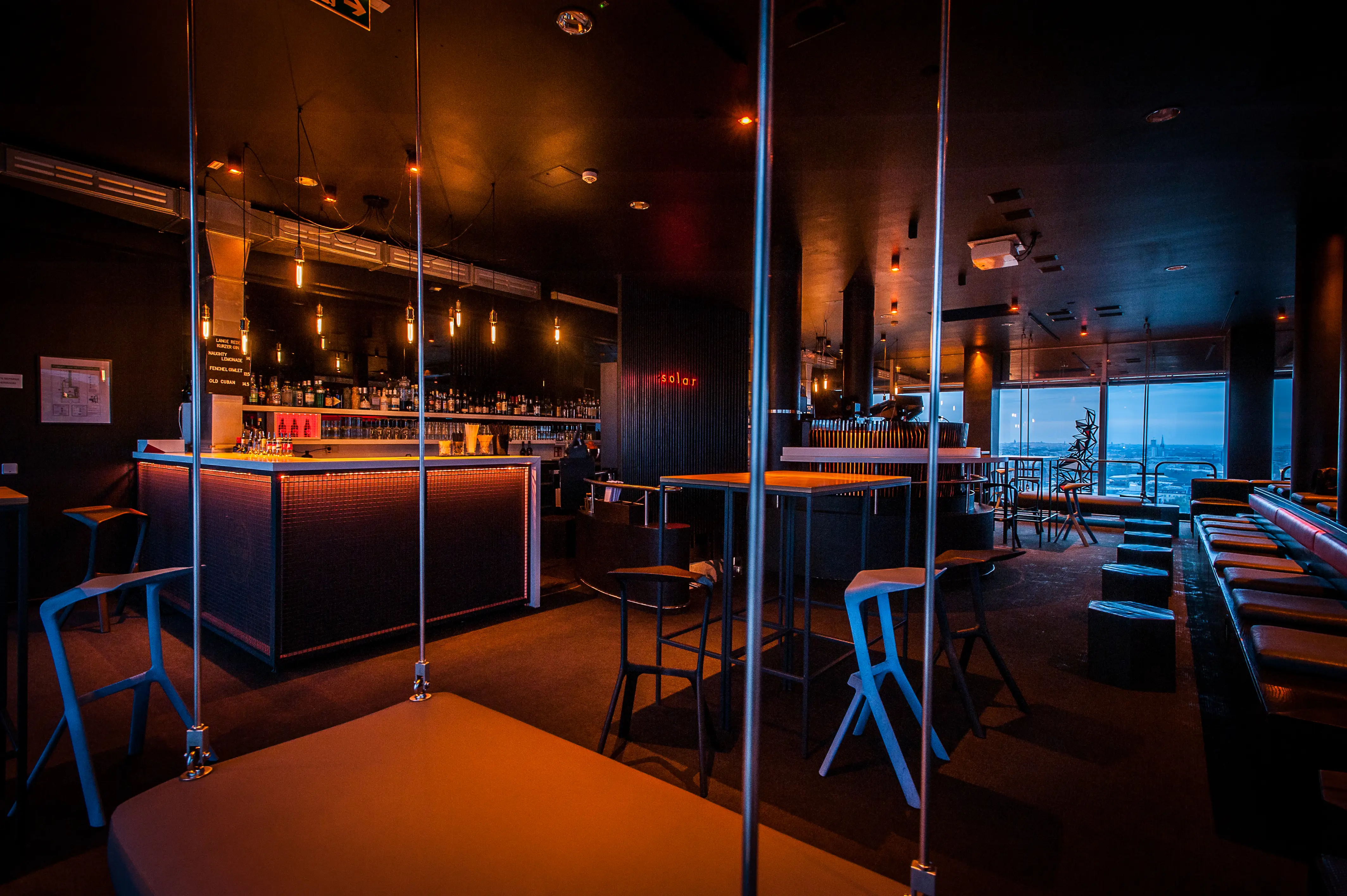 Dark bar room at SOLAR SKY LOUNGE with high tables and blue designer stools between floor-to-ceiling steel rods, the lit bar in the background and city windows along the side.