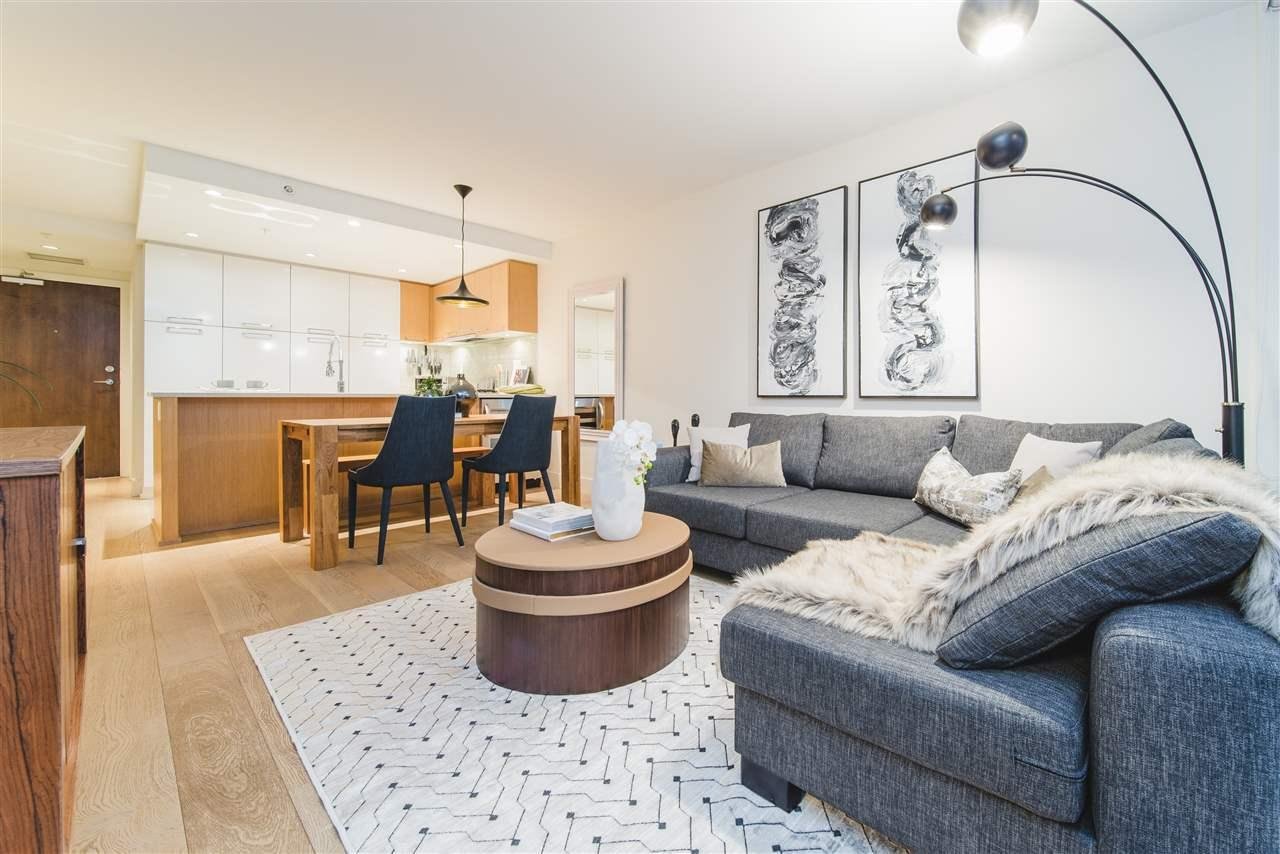 A cozy living room featuring a grey couch, round wooden coffee table, and a modern lamp, with a kitchen in the background.