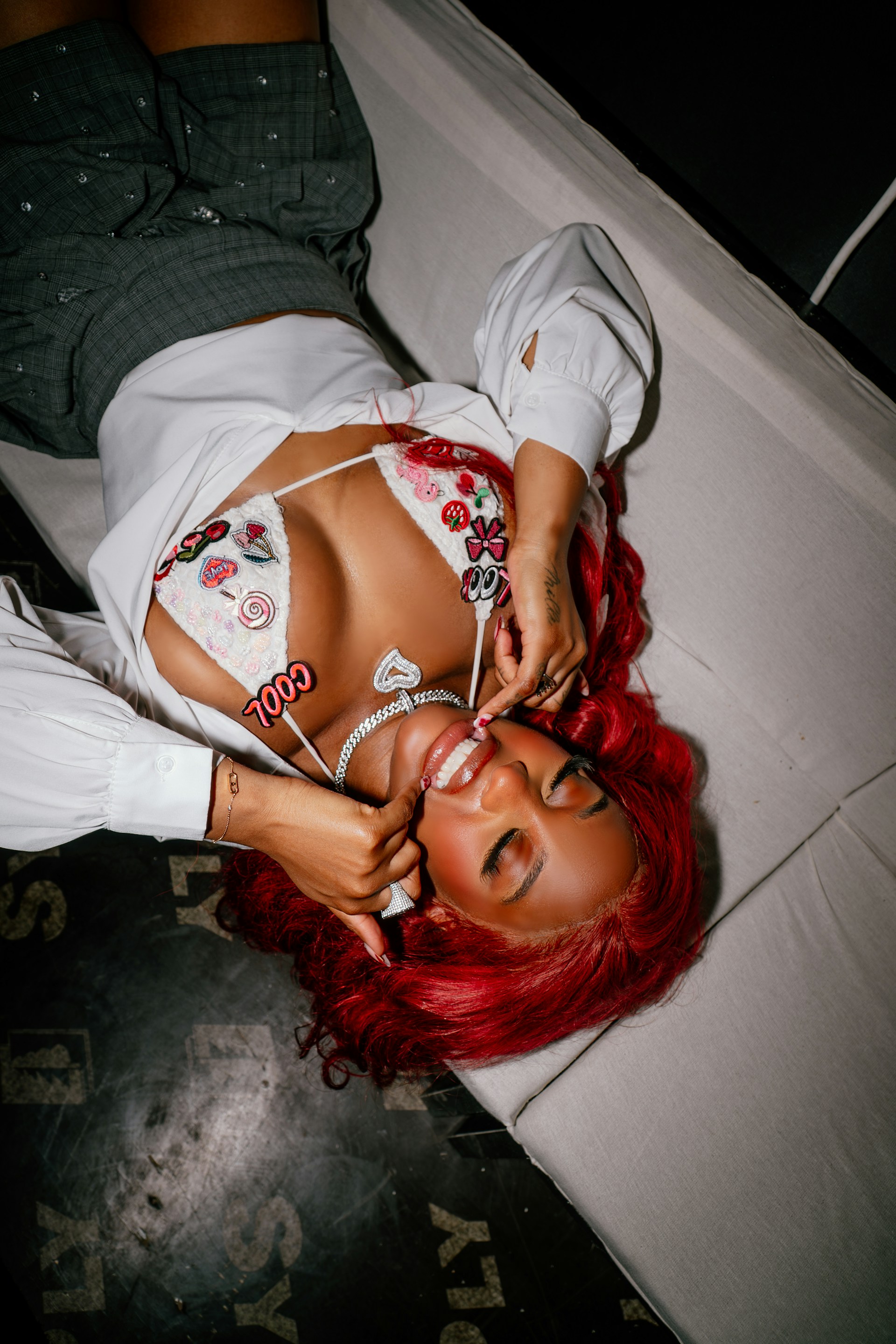 An overhead shot of the woman lying on a white surface. She is smiling and touching her teeth, highlighting her jewelry and the red tones of her hair.