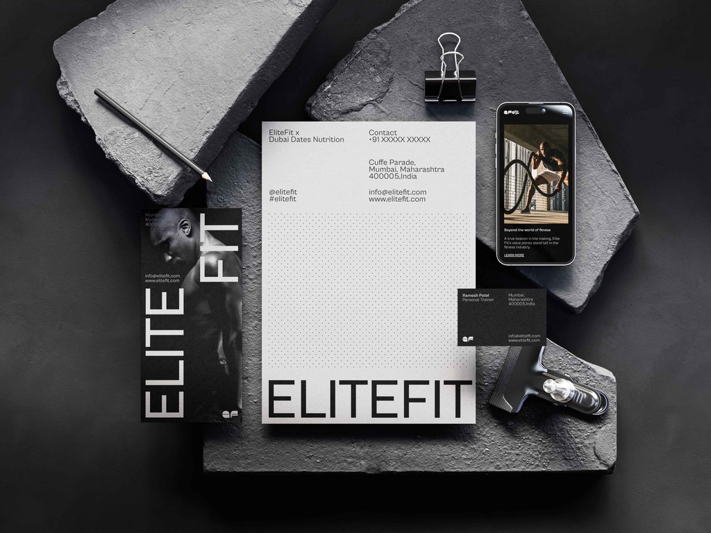 Flat lay of EliteFit branded items on a dark, textured surface, including a pencil, flyer, notepad, business card, smartphone, and massage gun, showcasing the EliteFit logo, contact information, and a muscular man working out.