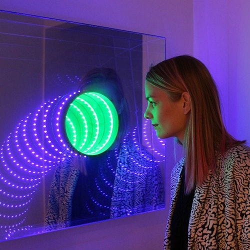 一位女士在一个镜面装置中仔细欣赏一件色彩斑斓的灯光艺术装置，装置中使用了绿色和紫色的LED灯。