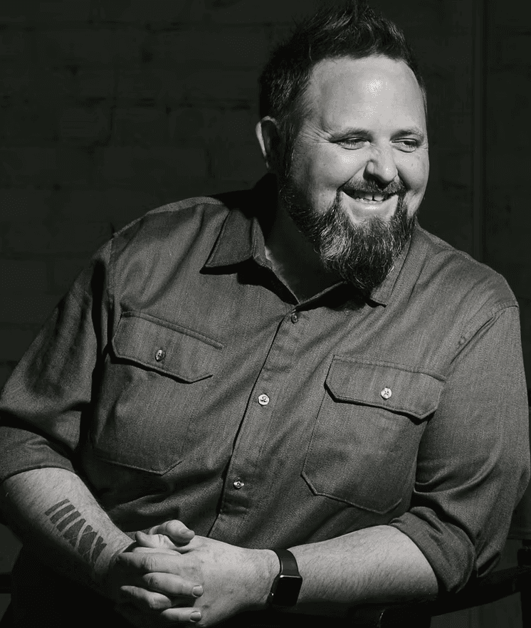 Director Kevin O’Brien, a specialist in branding and virtual production, sits smiling in a black-and-white portrait. He wears a button-down shirt with rolled sleeves, displaying a forearm tattoo and a smartwatch, radiating warmth and creative confidence.
