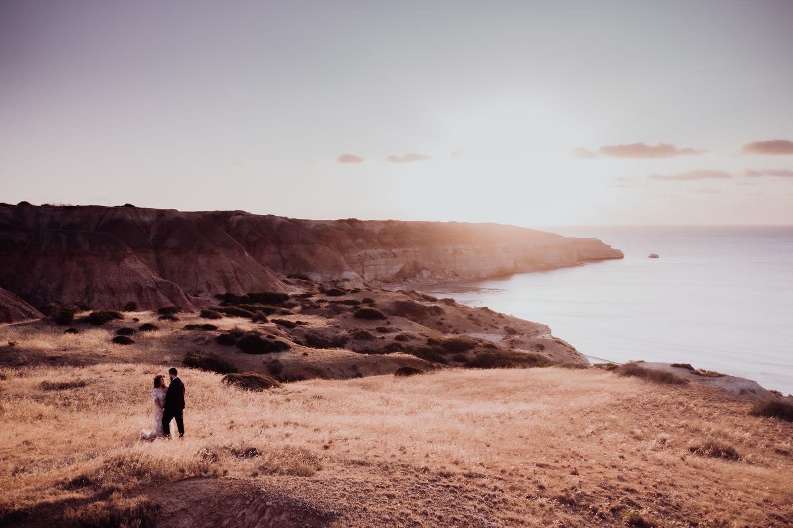 Two tiny figures on a grassy cliff above a rocky coast at sunset