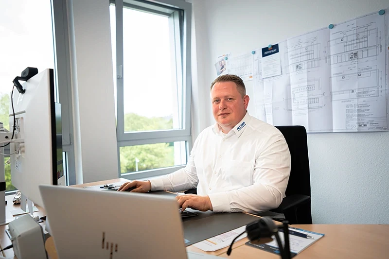 Projektleiter von RITTER Starkstromtechnik sitzt an seinem Schreibtisch im Büro und arbeitet am Computer. Im Hintergrund sind technische Pläne an der Wand angebracht.