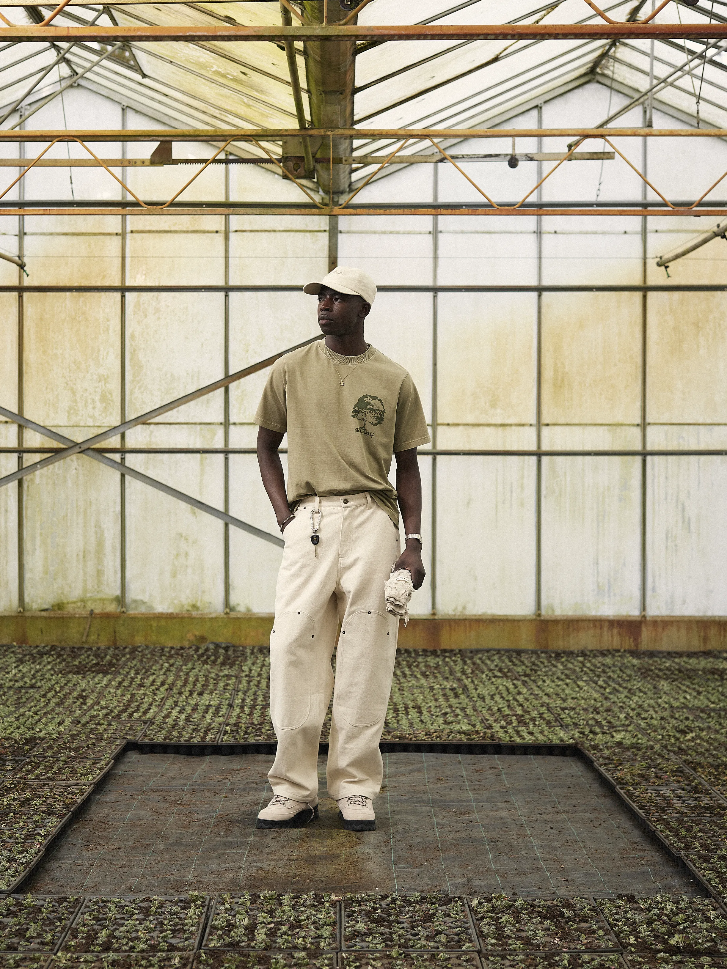Dans une serre, le mannequin porte un pantalon large beige, un t-shirt vert clair et une casquette "Fuck off I'm gardening"