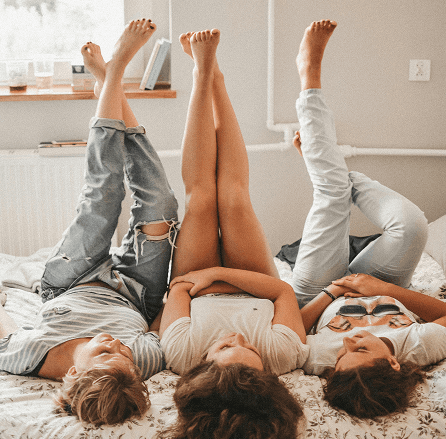 Three friends lying on a bed with their legs up against the wall, laughing and enjoying a relaxed moment together.