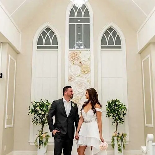 Ett par, klädda i bröllopskläder, ler och håller varandra i händerna i ett ljust kapell med blommiga dekorationer och välvda fönster.