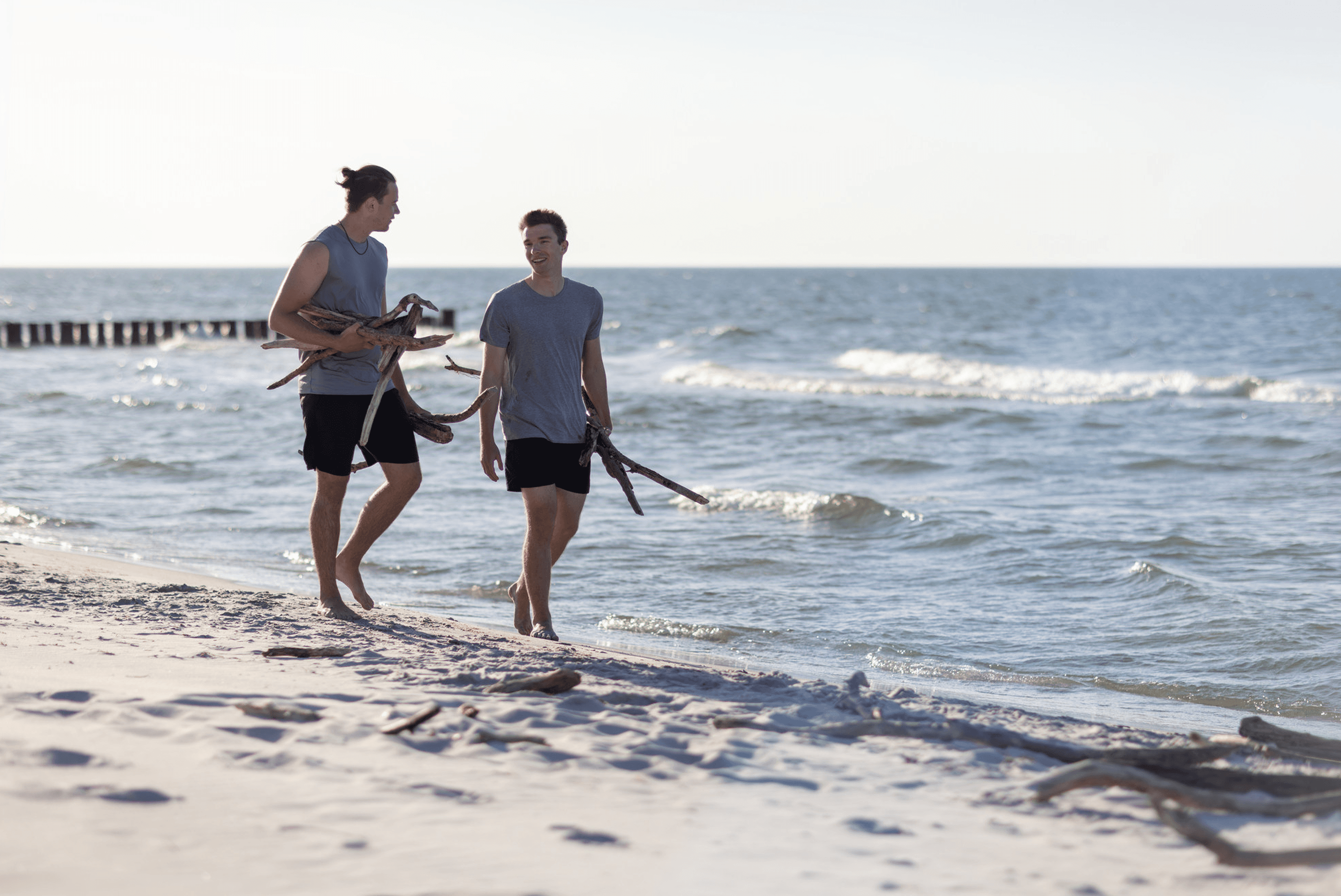 Two men walk on a beach carrying driftwood.
