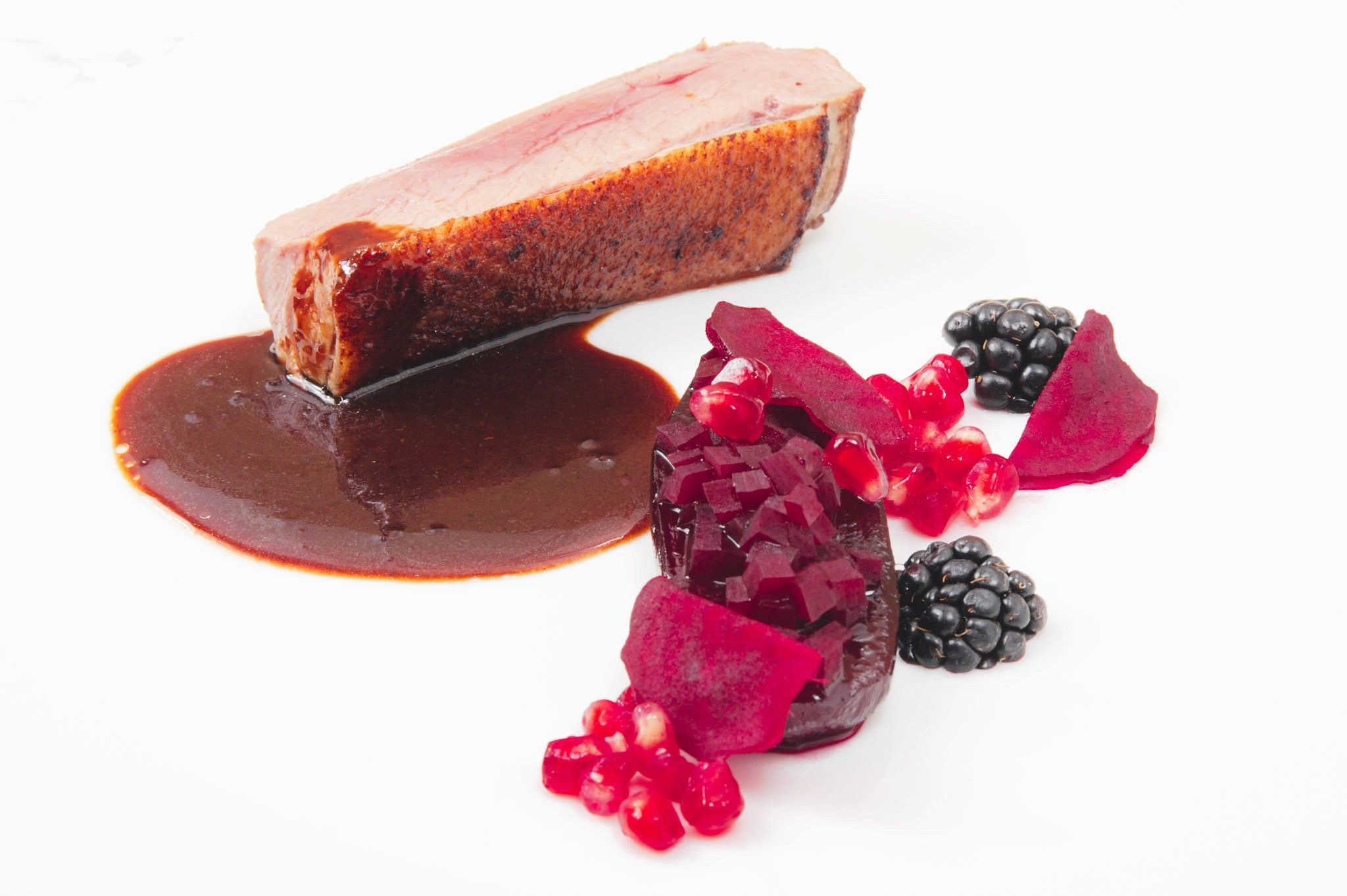Close shot of duck breast with blackberry sauce beside glazed beetroot, pickled beet slices, blackberries, and pomegranate.