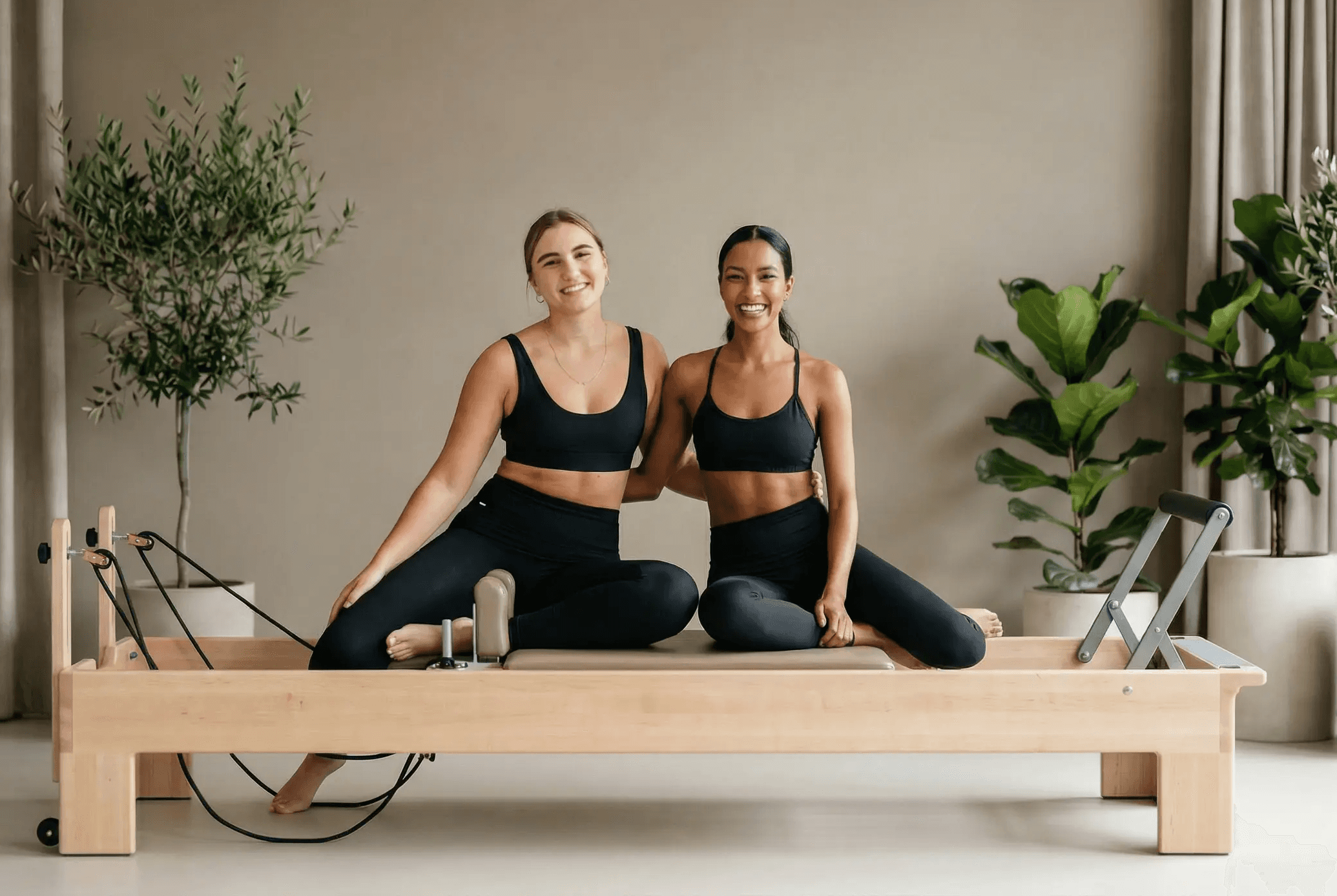 Two women sitting together on a reformer machine next to each other smiling at camera
