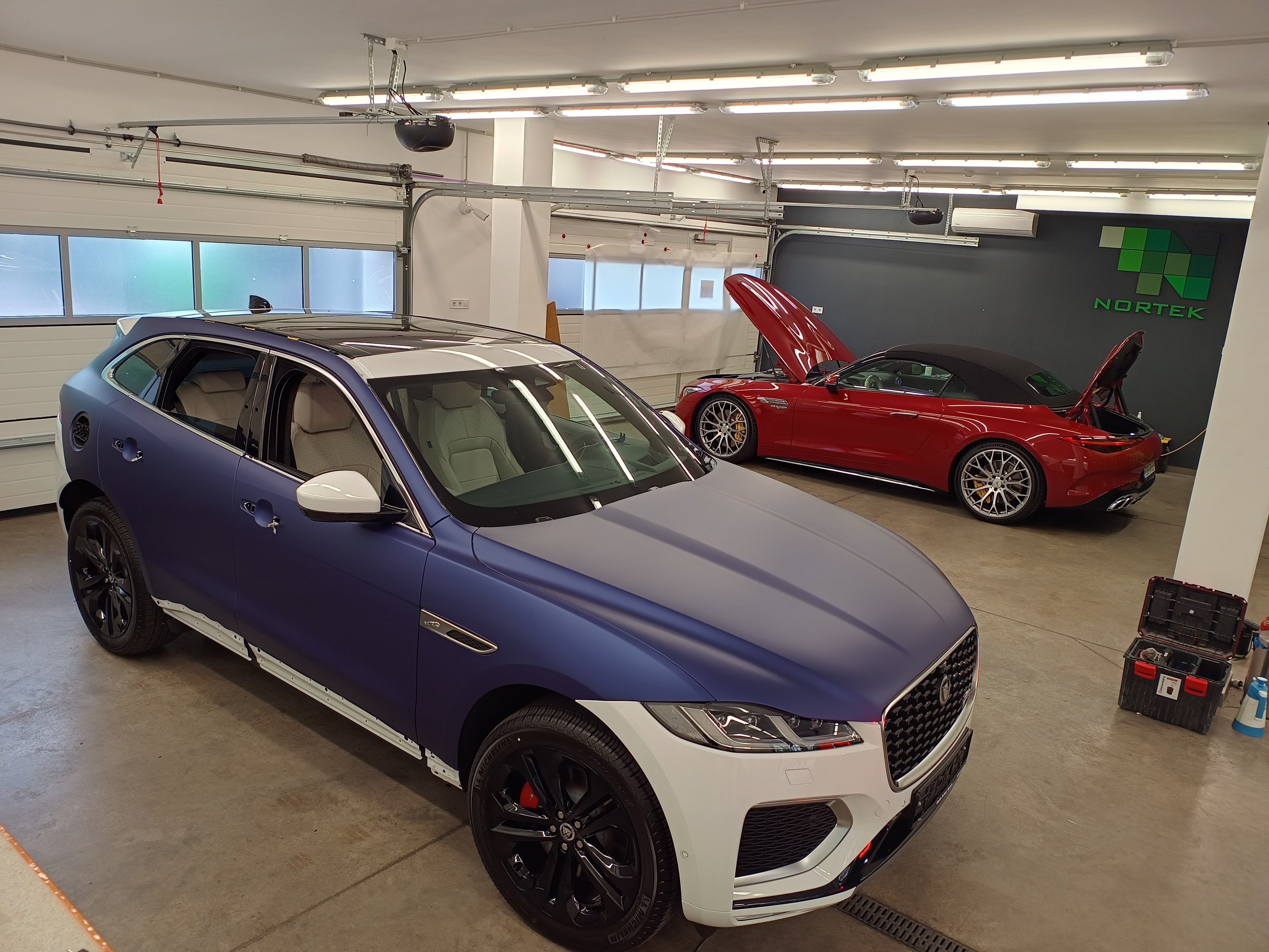 Matt sötétkék prémium autófóliával (wrap) átszínezett Jaguar luxus SUV a Nortek budapesti műhelyében, a háttérben egy nyitott motorháztetejű, fényes piros Mercedes SL63 AMG Cabrio sportautóval.