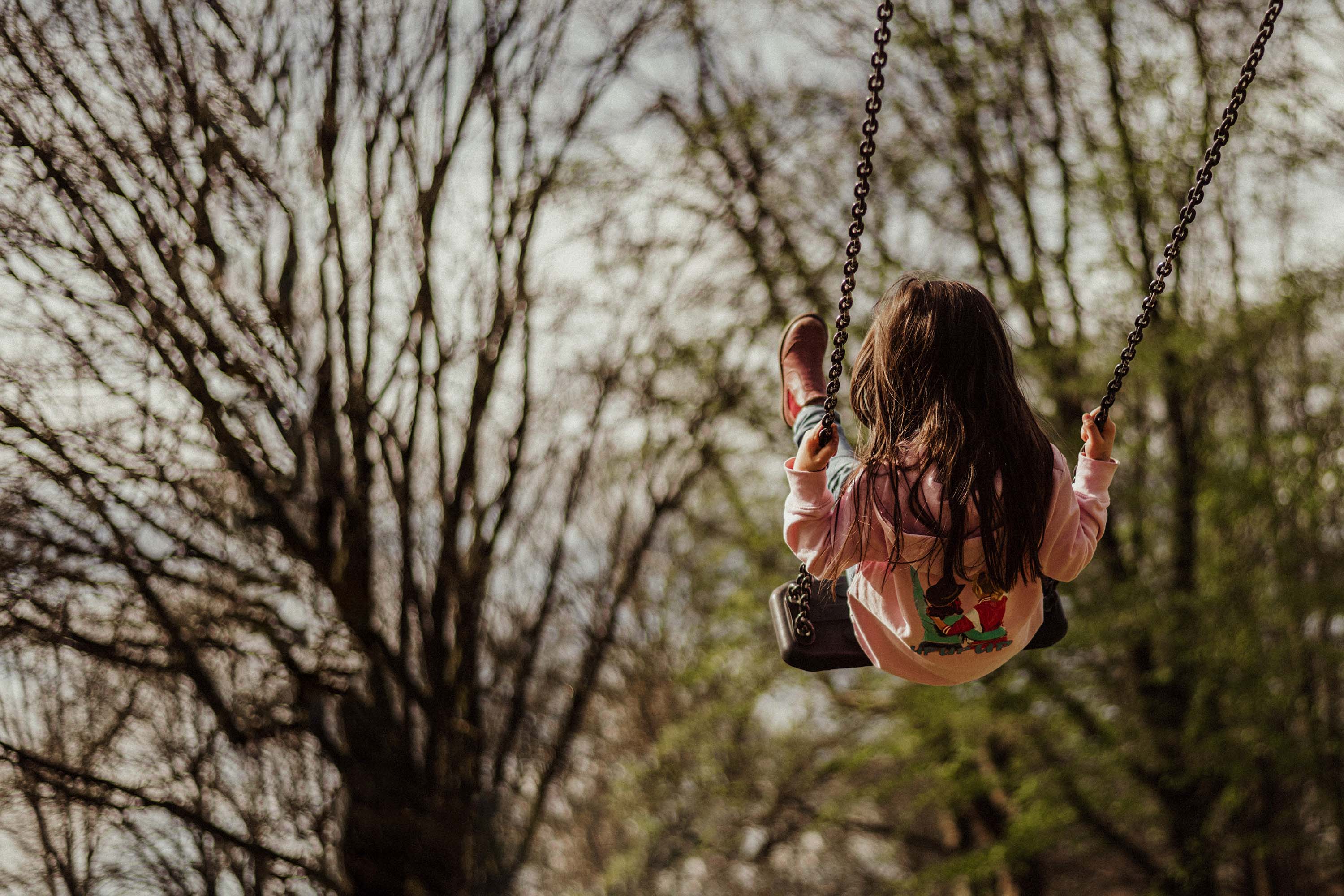 Authentische Kindergartenfotografie Gevelsberg: Ein Mädchen auf einer Schaukel schwingt hoch in die Luft, fotografiert von PK photography 4 Kids NRW. Der Hintergrund zeigt unscharfe Bäume und ein weiches Bokeh