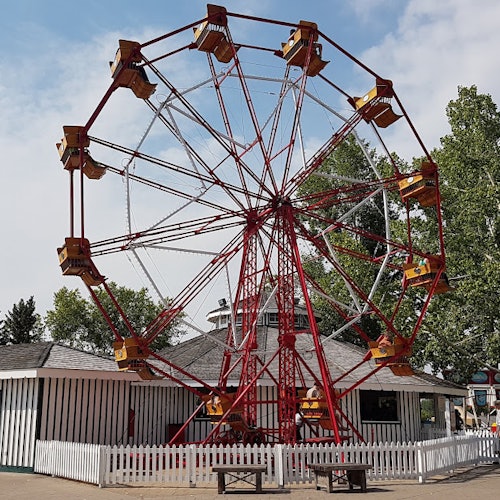 Roda-gigante vermelha e amarela em um parque de diversões, cercada por uma cerca branca, com árvores e um edifício ao fundo.