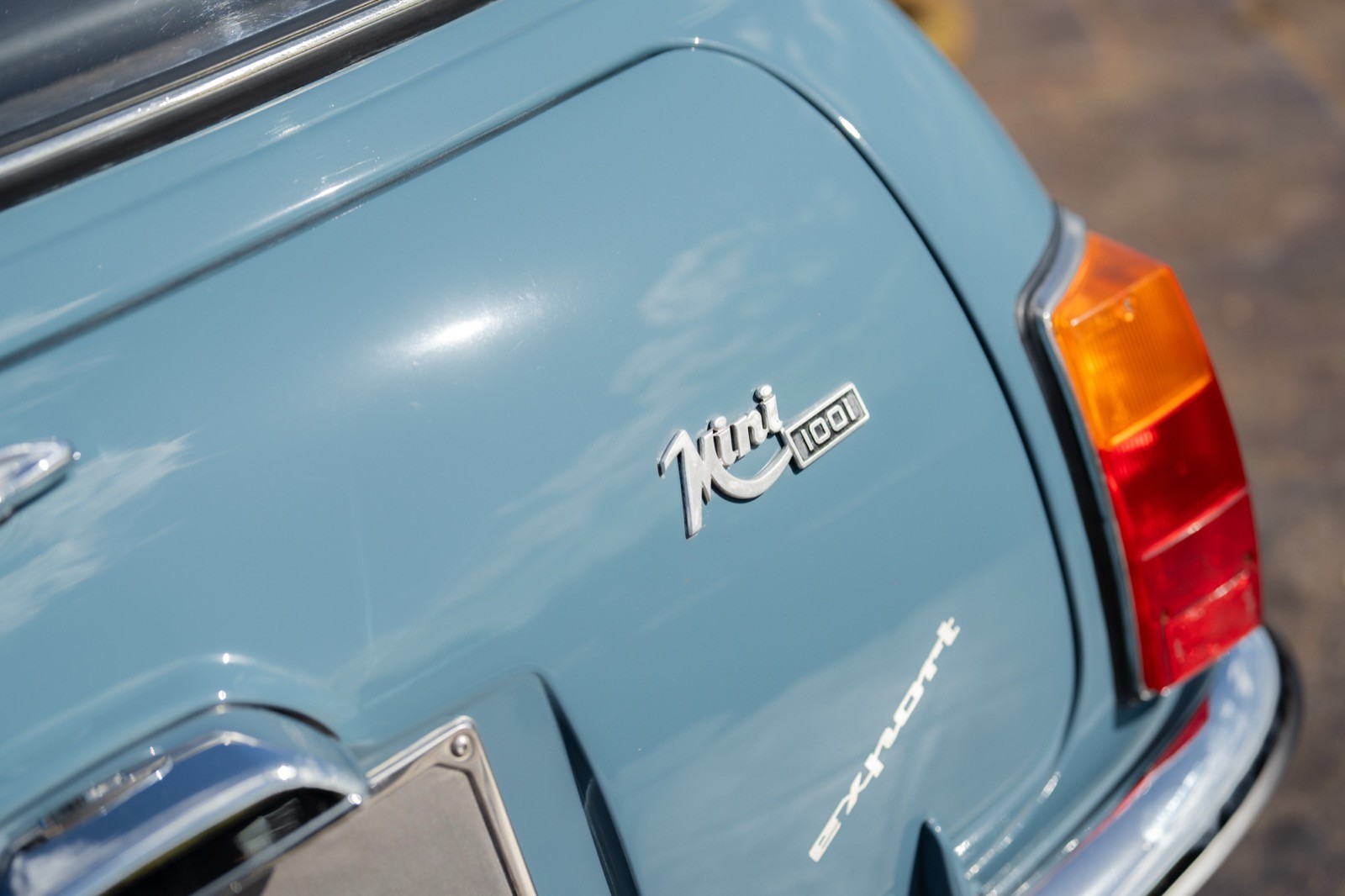 Close-up of the rear of a light blue vintage car, showcasing its emblem and orange taillight.