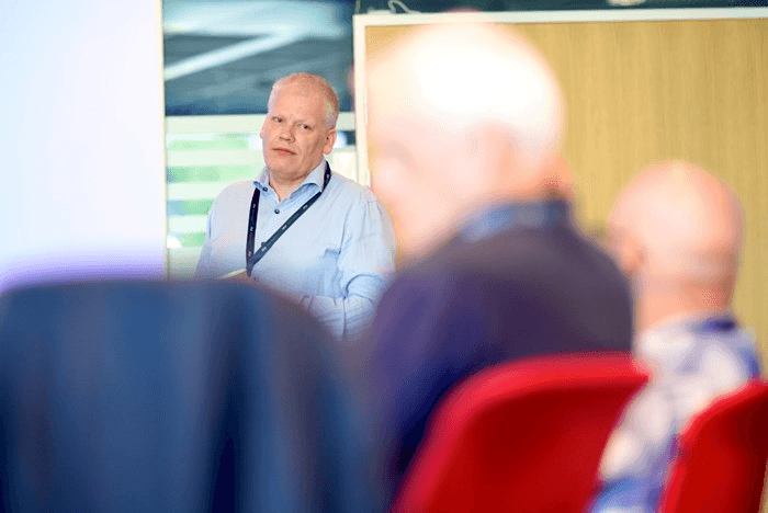 A speaker presents to an audience, with attendees seated in blurred focus in the foreground.