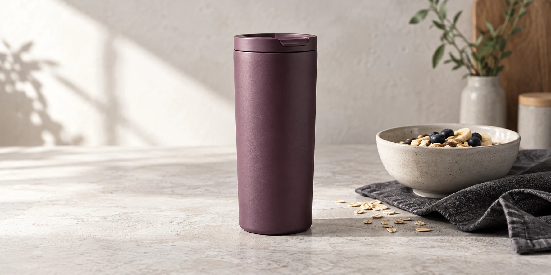 Muted plum travel tumbler on a light stone table beside a breakfast bowl and charcoal linen