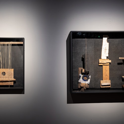 Dalawang shadow box na may itim na frame ang nagtatampok ng mga assemblage ng nahanap na mga bagay at piraso ng kahoy na nakapaskil sa isang kulay abong dingding.