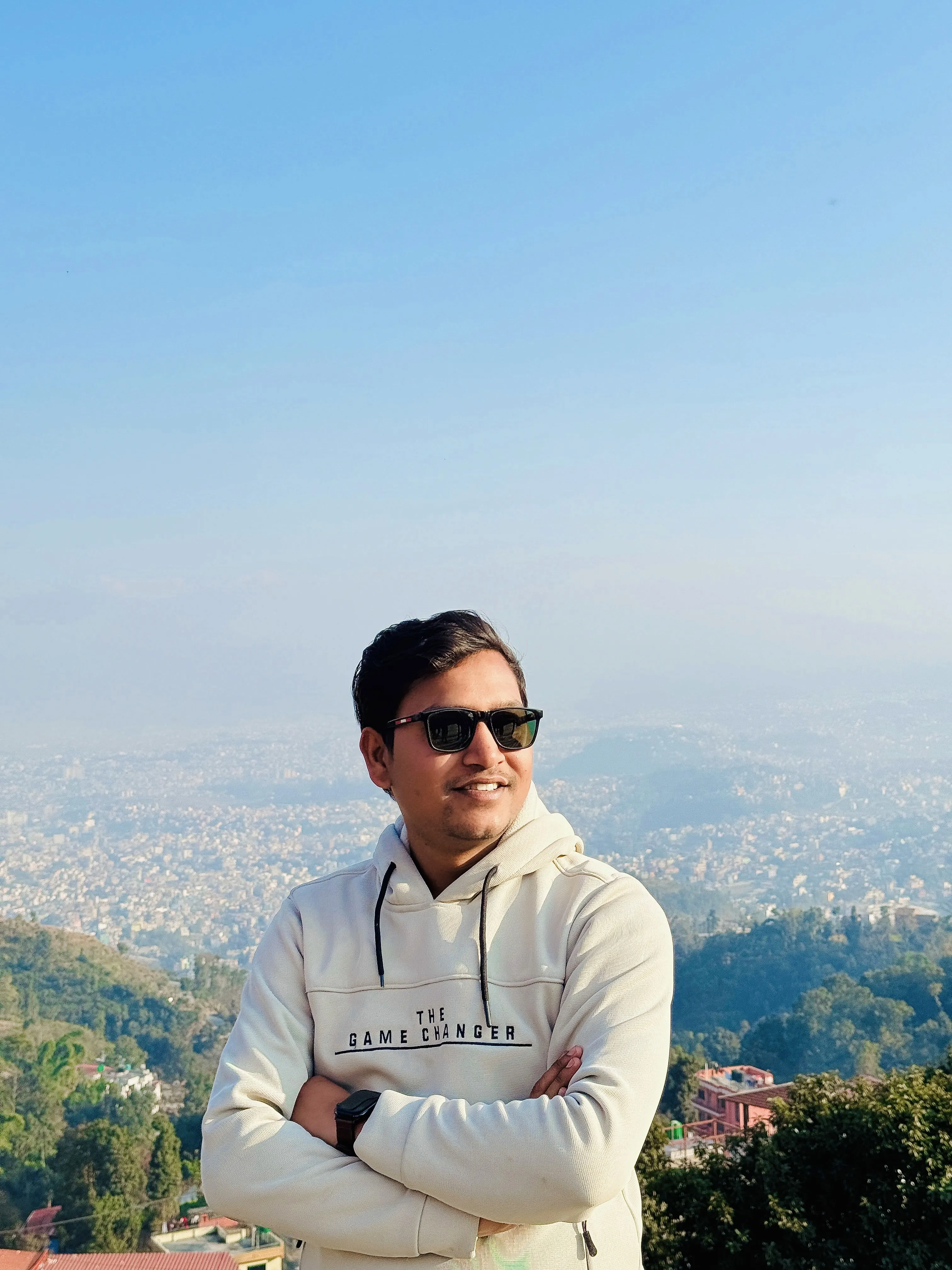 Man in sunglasses and hoodie with arms crossed overlooking city.