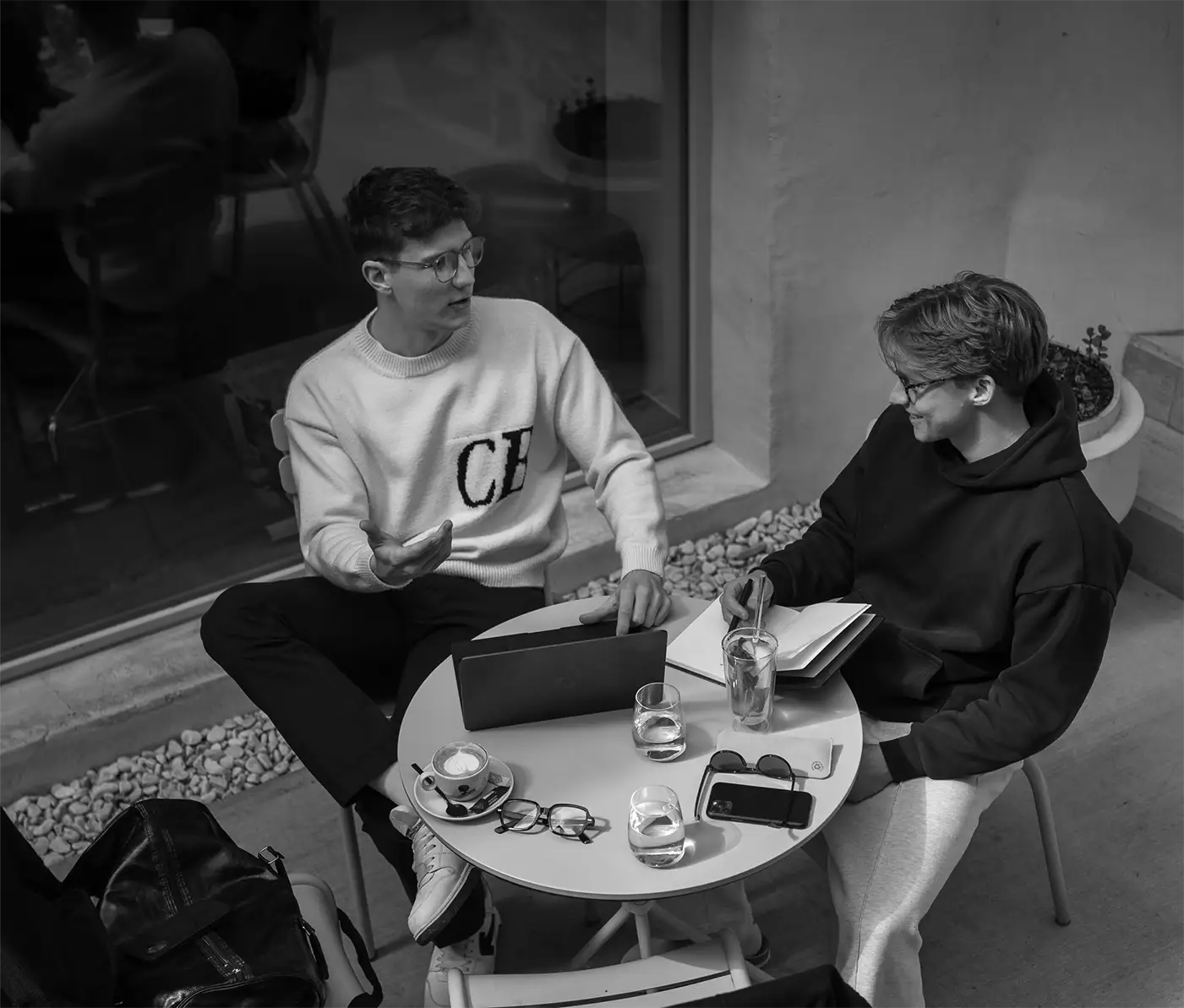 Bild in schwarz weiß von Felix Maier und Paul Schlager im Gespräch an einem Café-Tisch mit Laptop, Notizbuch und Getränken – lockerer Austausch über Design- oder Marketingstrategien im modernen Arbeitsumfeld.