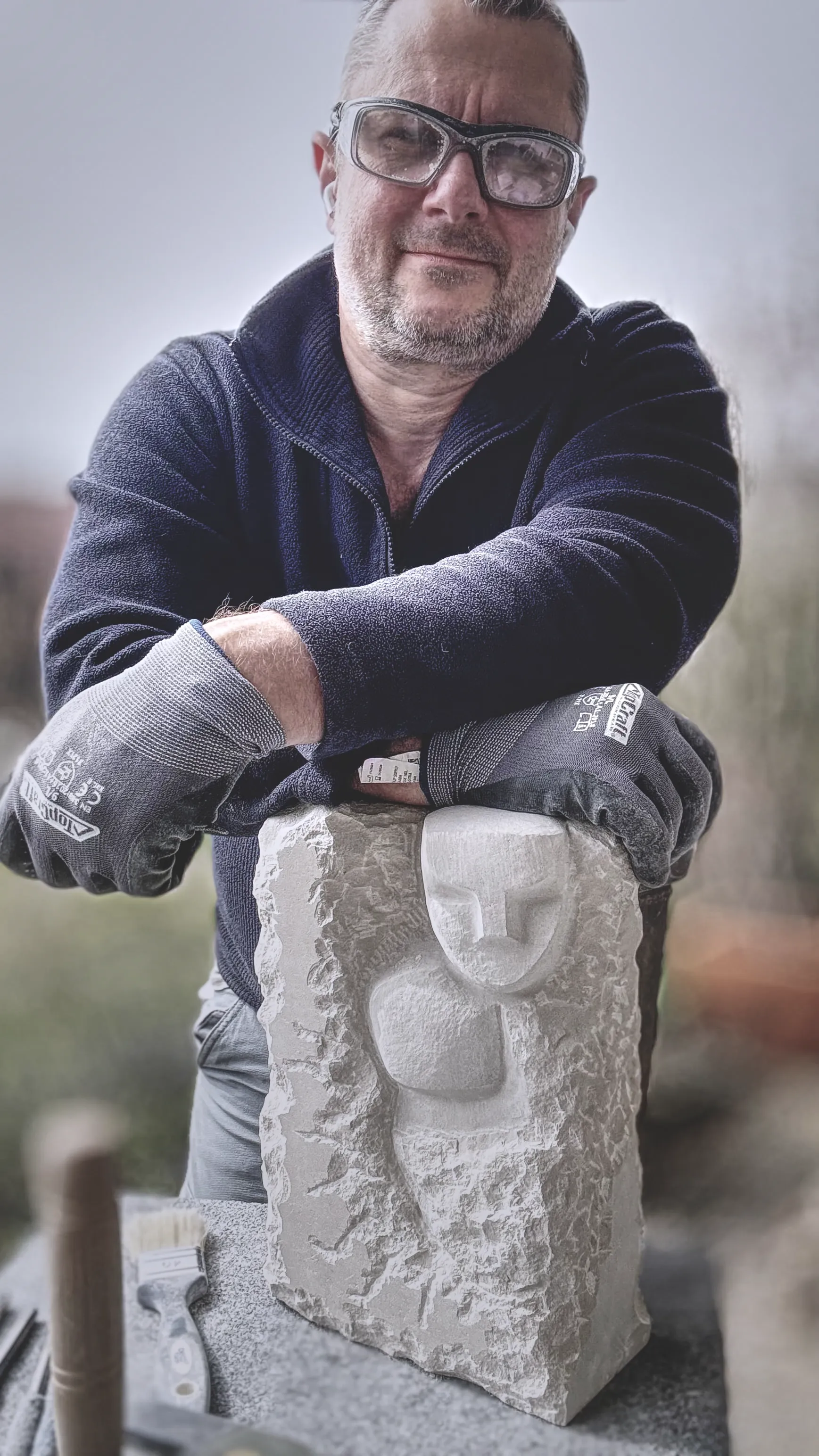 Bildhauer mit Schutzbrille und Arbeitshandschuhen lehnt sich lächelnd auf eine frisch gemeißelte, helle Steinskulptur im Außenbereich