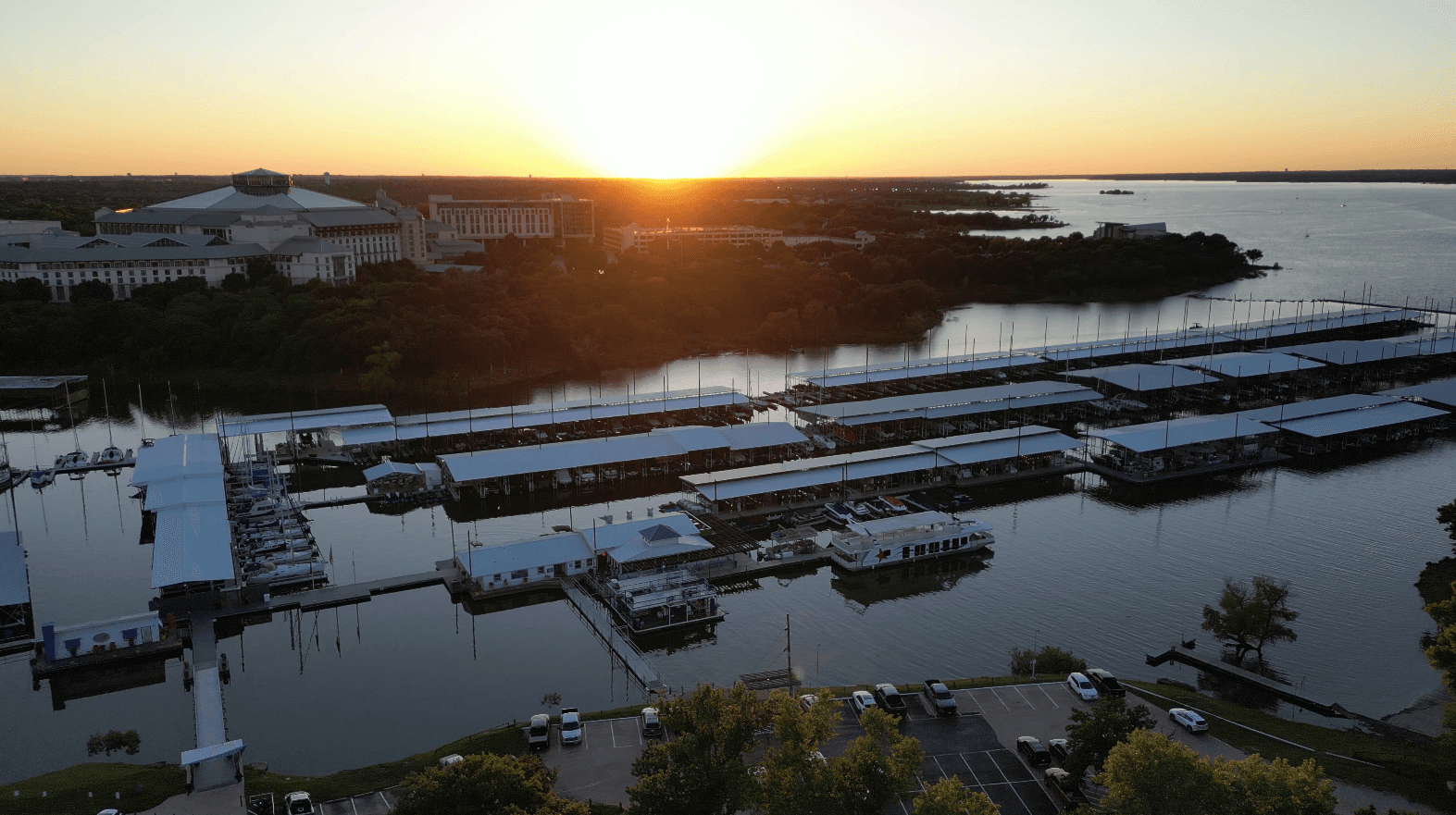 A scenic aerial view of a marina at sunset, featuring rows of boats docked at covered piers, surrounded by calm waters and lush greenery, with a glowing horizon in the background.