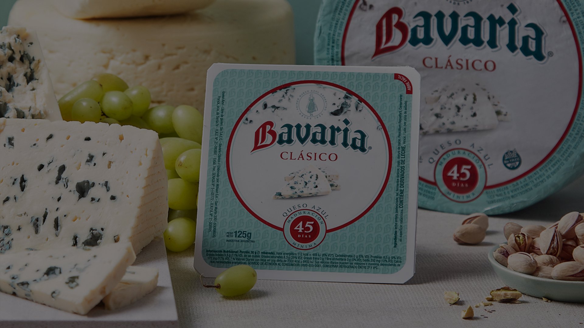 Packaging and slices of Bavaria Clásico blue cheese are surrounded by fresh green grapes and a small pile of pistachios on a table. The scene conveys a gourmet and inviting atmosphere, highlighting the cheese's 45-year tradition.