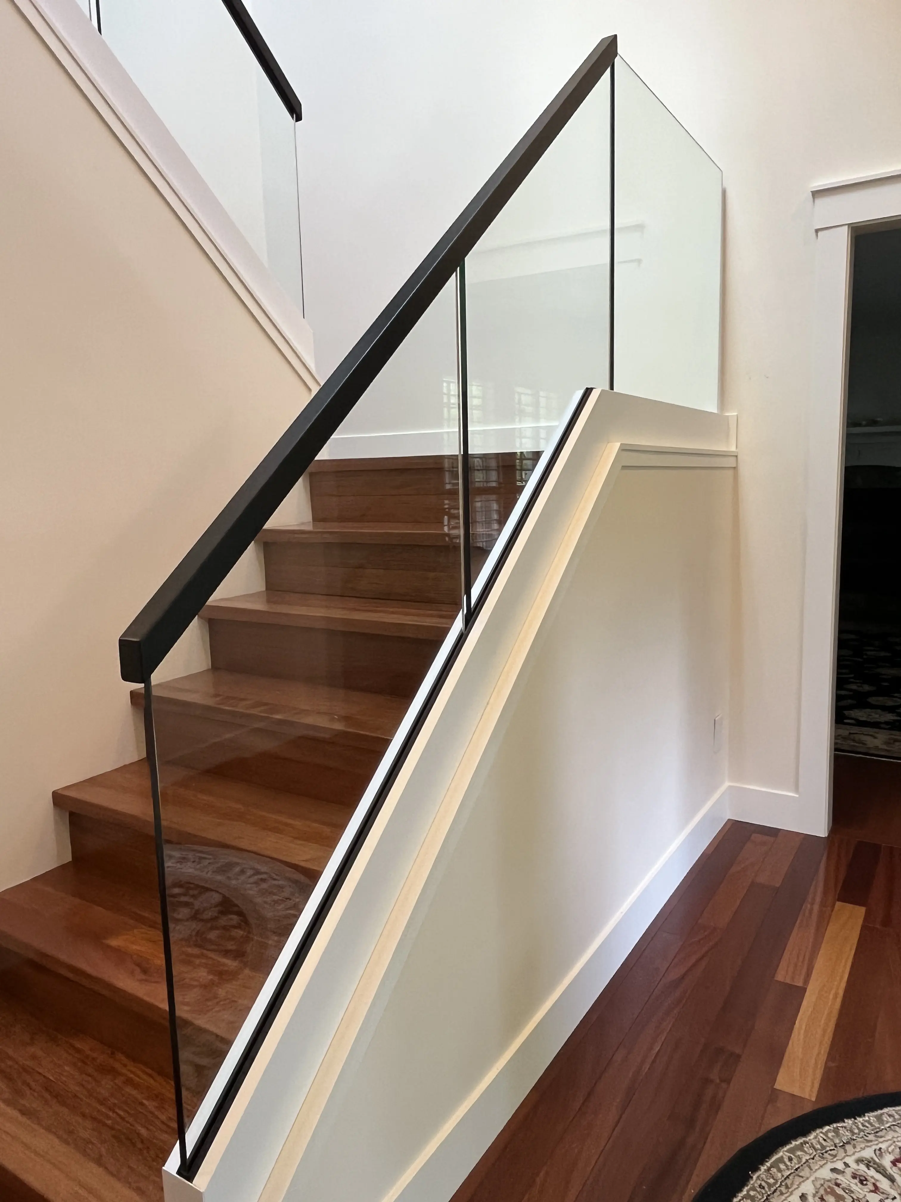 An interior staircase with wood treads and a dark diagonal wood handrail, paired with a frameless glass guard that rises directly from a white painted stringer cap along the stair edge, with no visible surface-mounted hardware interrupting the glass. Warm hardwood flooring runs along the lower level, with a doorway and area rug visible through the opening beside the stairs.