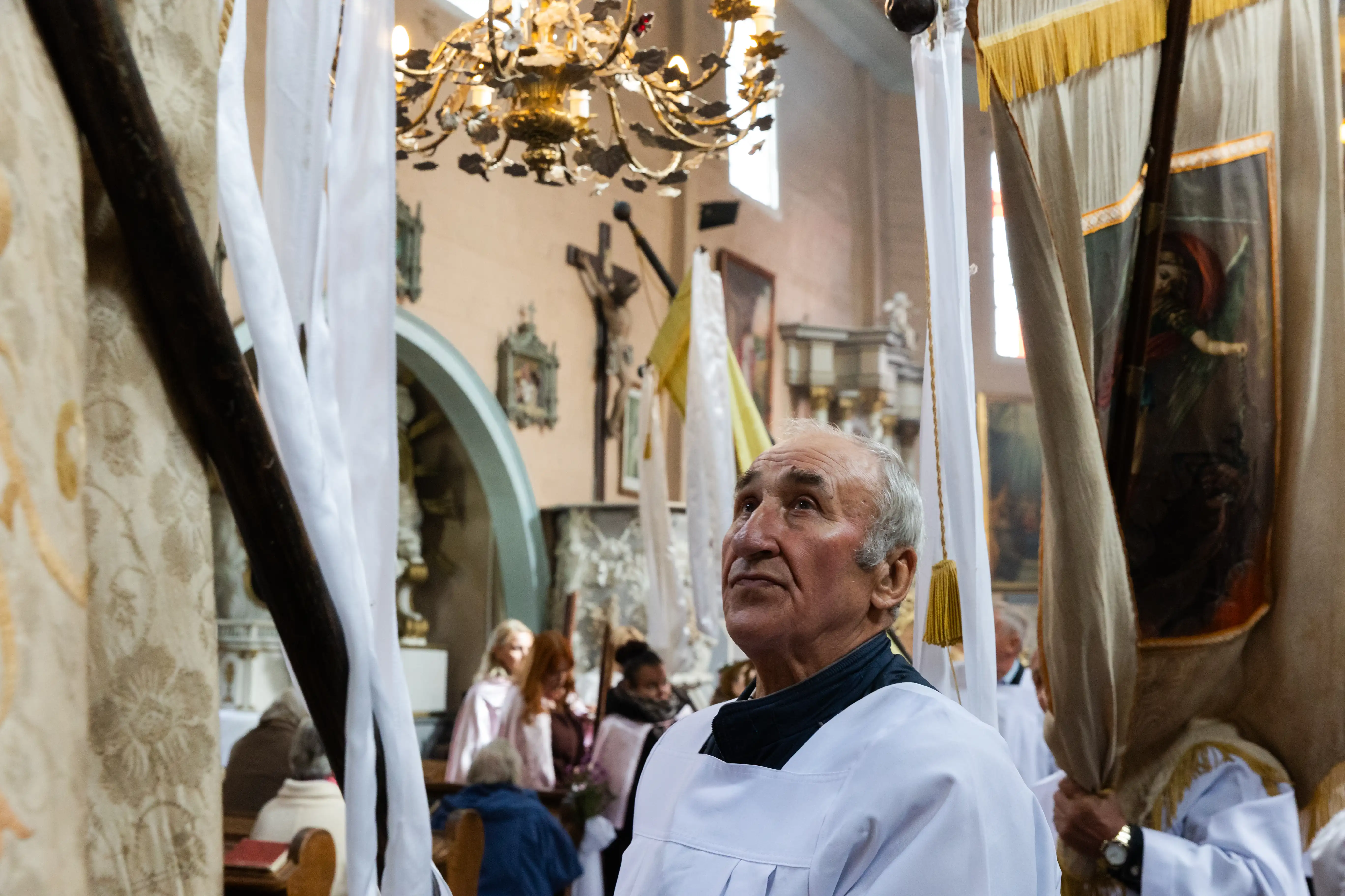 Mężczyzna w białym stroju liturgicznym patrzy w górę podczas ceremonii w kościele. W tle widoczni uczestnicy z sztandarami, a sufit zdobi żyrandol. Nastrojowa atmosfera w otoczeniu religijnych symboli.