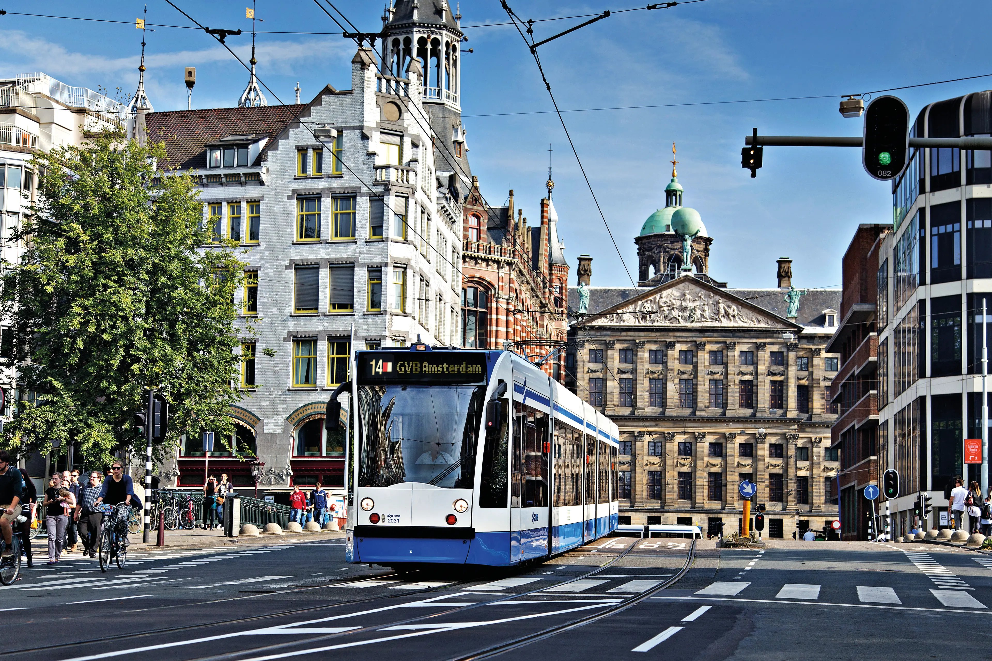 Tram van GVB Amsterdam, lijn 14, rijdt door een historische straat met karakteristieke gebouwen op de achtergrond. De afbeelding toont de combinatie van modern openbaar vervoer en de charmante architectuur van Amsterdam, met een heldere blauwe lucht en levendige straatactiviteit. Deze foto benadrukt de toegankelijkheid van de stad en het gebruik van trams als een populaire manier van vervoer in Amsterdam, ideaal voor toeristen en bewoners.