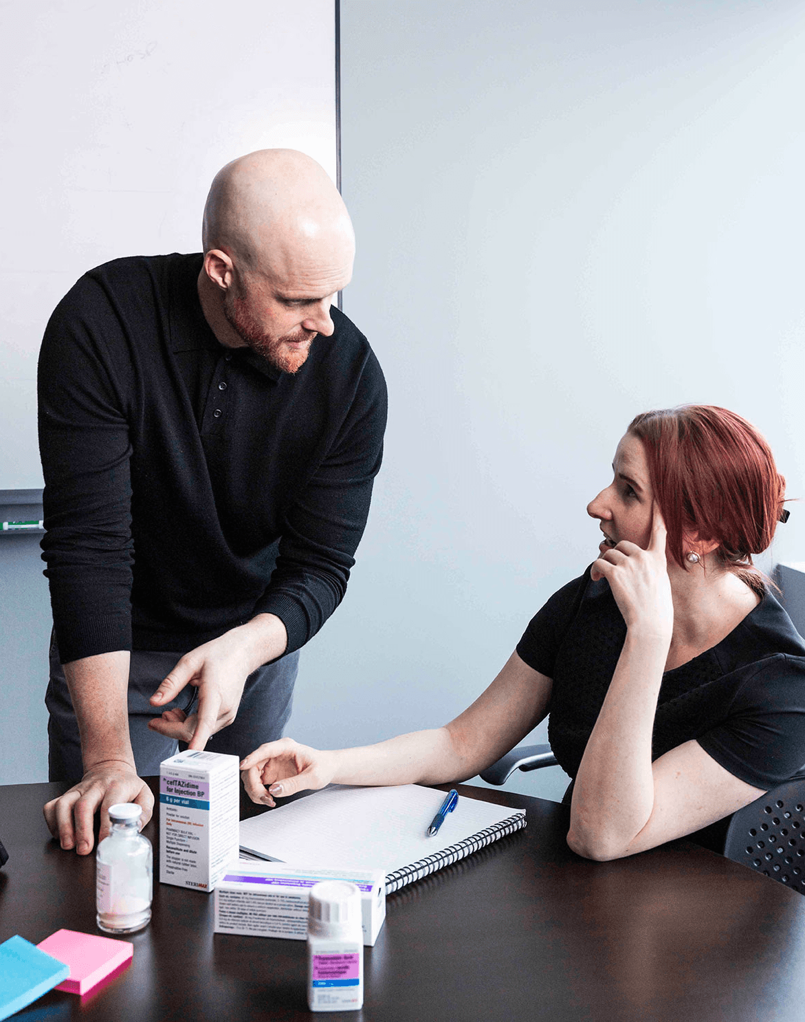 Two colleagues have a discussion at an office table with notes and pill bottles.