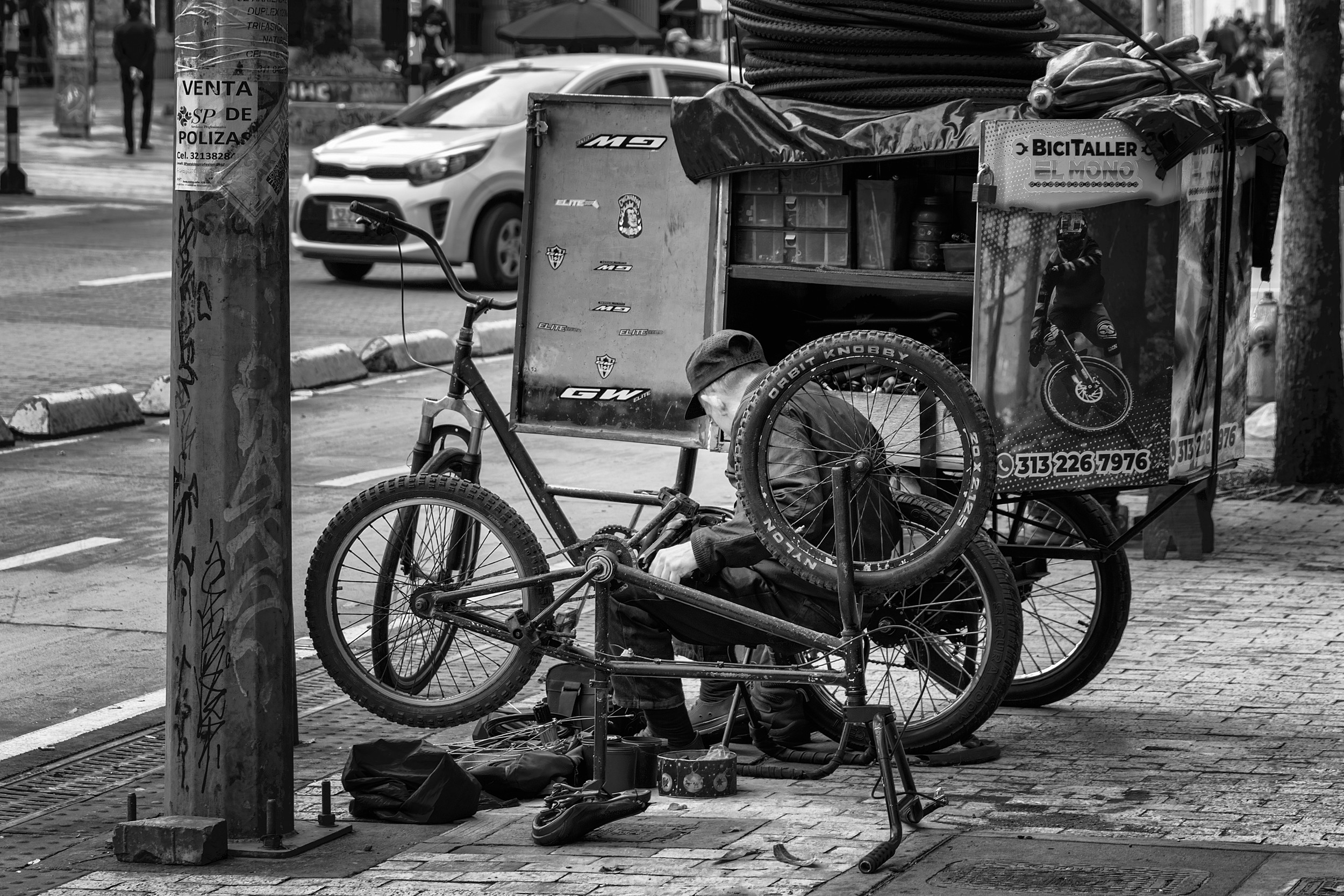 A bicycle repairman works crouched beside his mobile workshop cart on a Bogotá sidewalk, surrounded by spare tires, tools, and an upside-down bike.