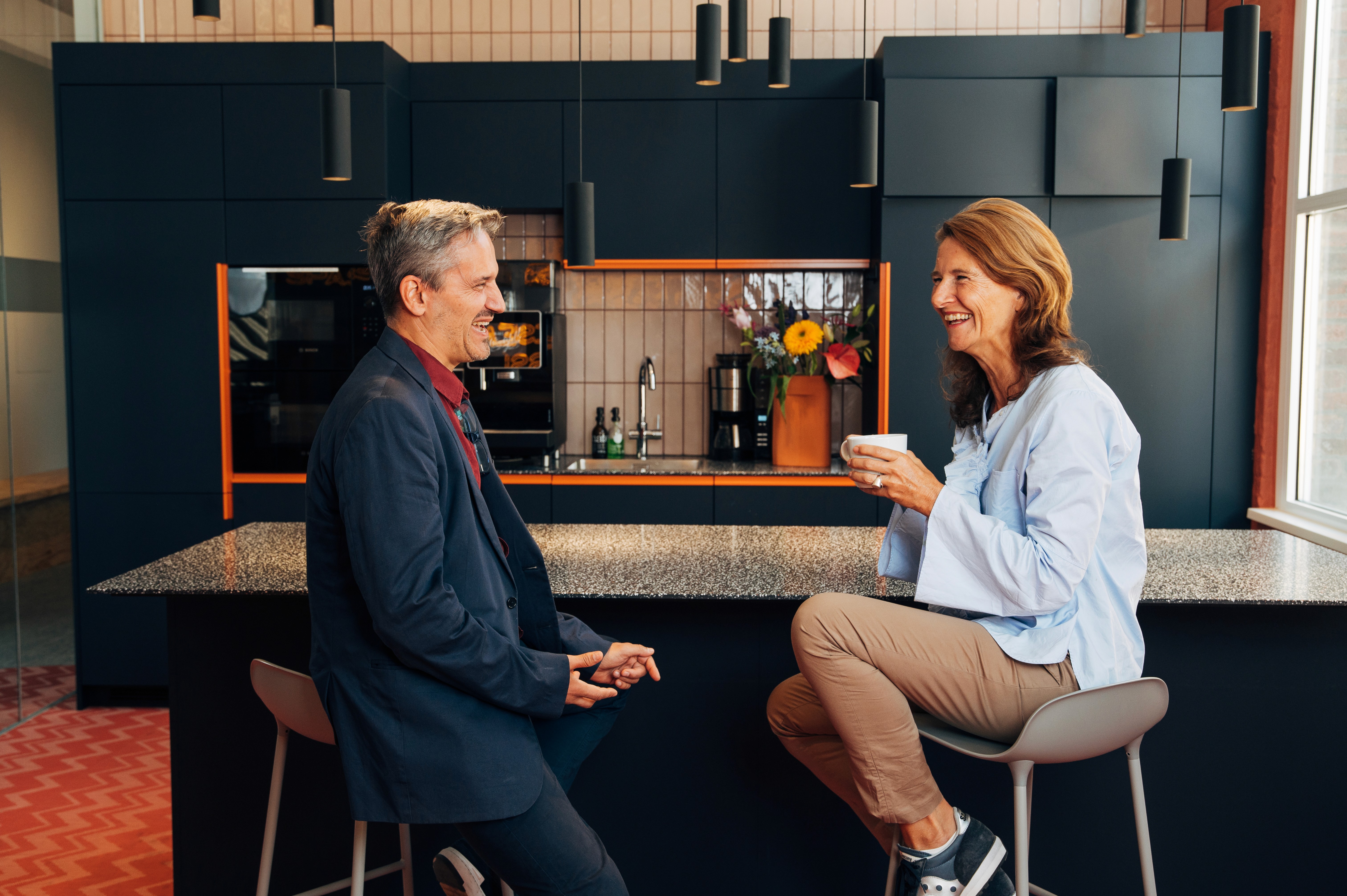 Two people are engaged in a conversation at a kitchen counter, with colorful items in the background.