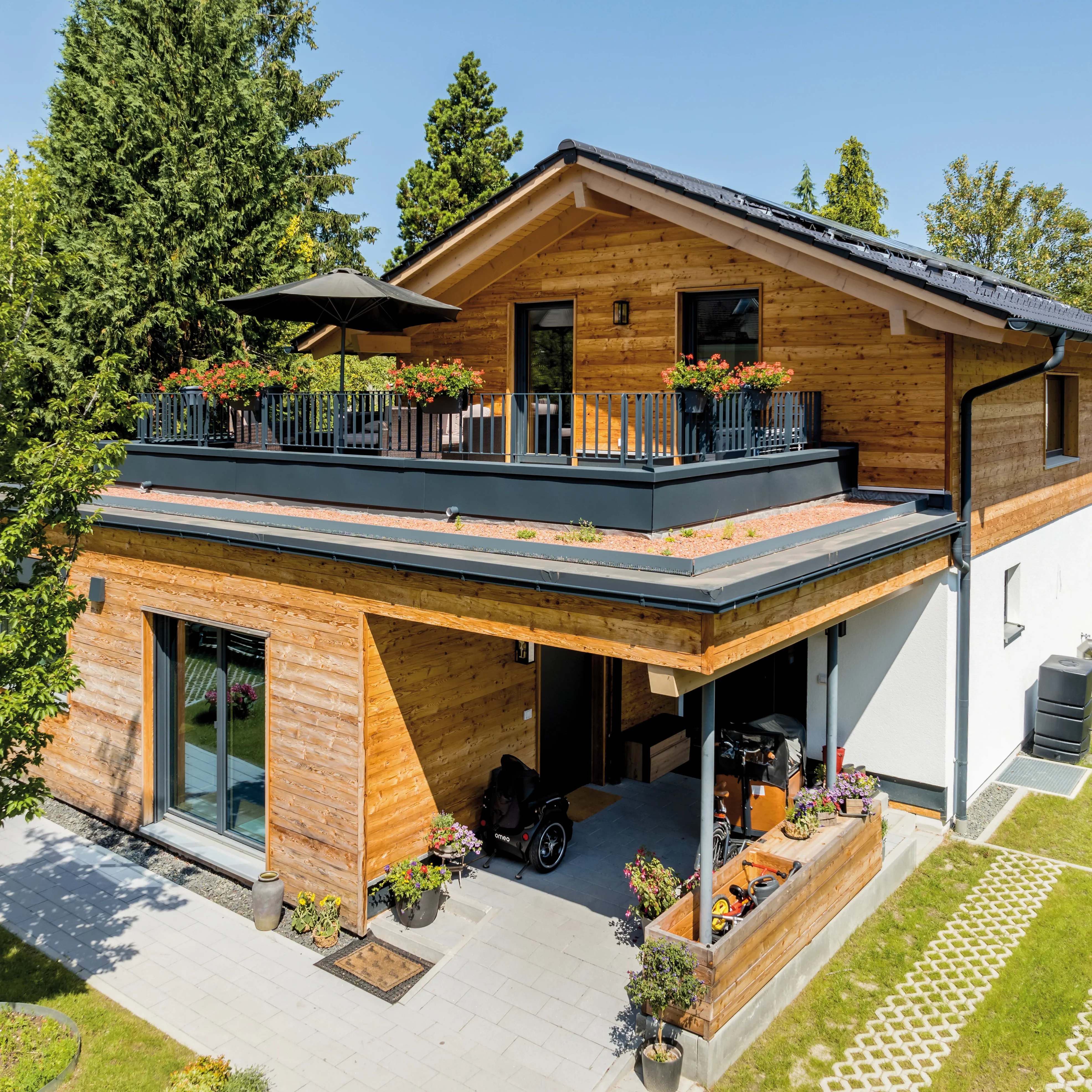 Modernes Holzhaus mit großzügigem Balkon, überdachtem Parkplatz und gepflegter Außenanlage, sonniger Tag.