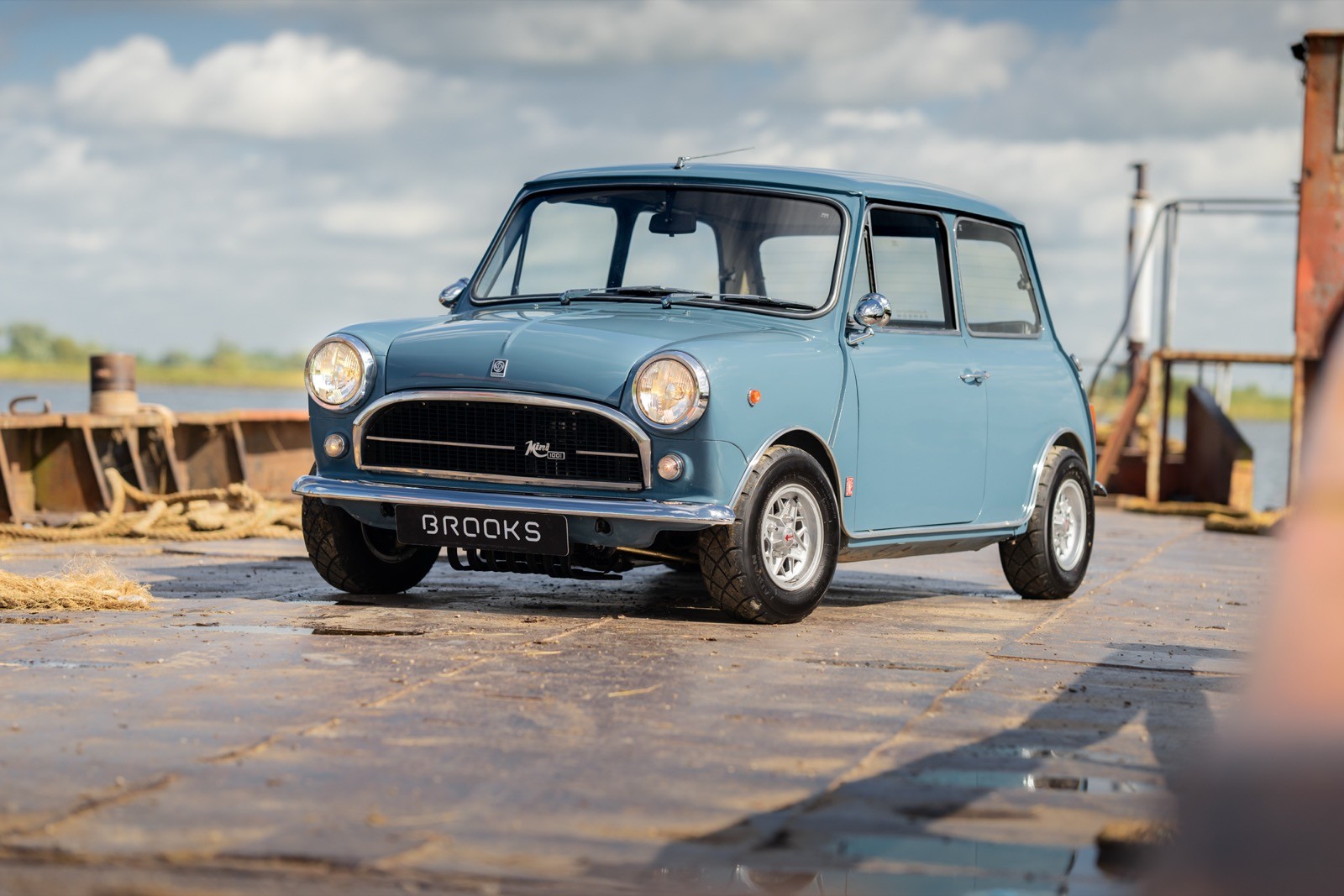 A vintage blue car parked on a wet road, with fields and a cloudy sky in the background.