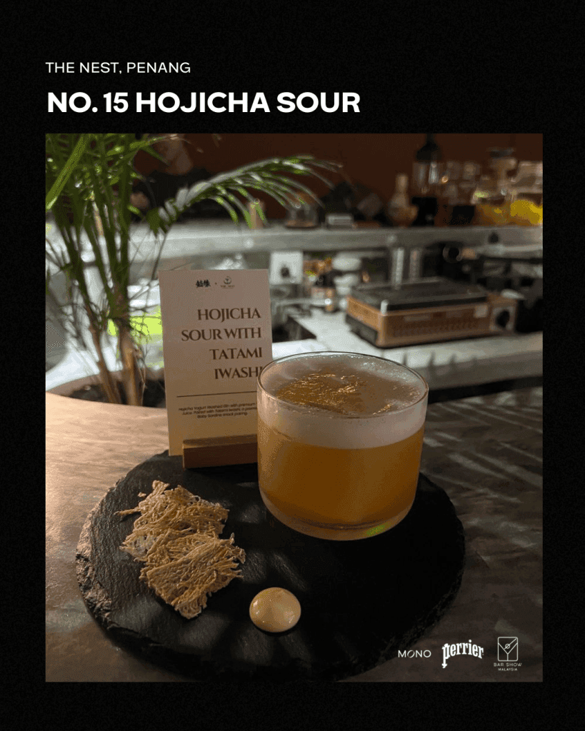 A glass of Hojicha Sour cocktail served on a dark stone plate, accompanied by tatami iwashi and a small white garnish. The background shows a bar setting with greenery.