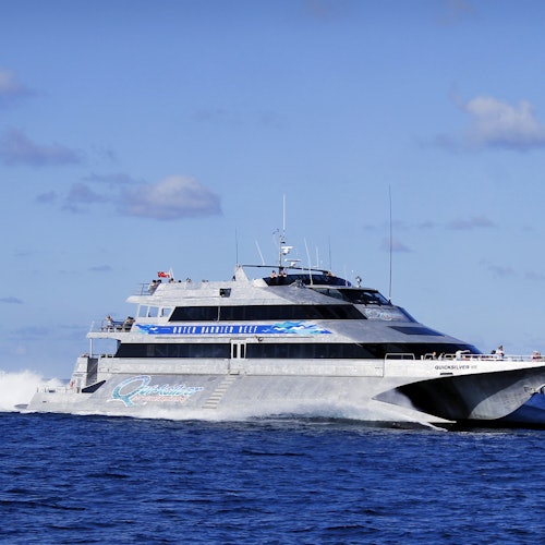A large, modern catamaran speeding through the ocean on a clear day, with a blue sky and a few clouds in the background.