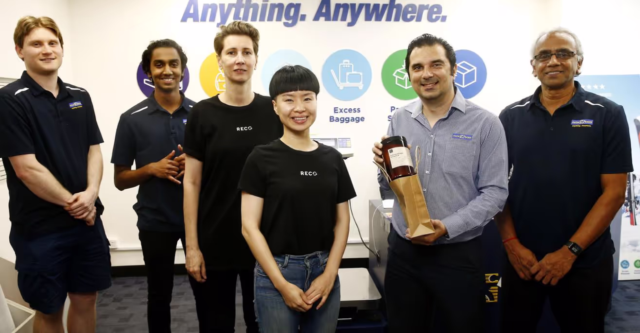 Six people standing in front of a PACK & SEND banner reading Anything. Anywhere., two of them in ReCo t-shirts, one holding a packaged bottle