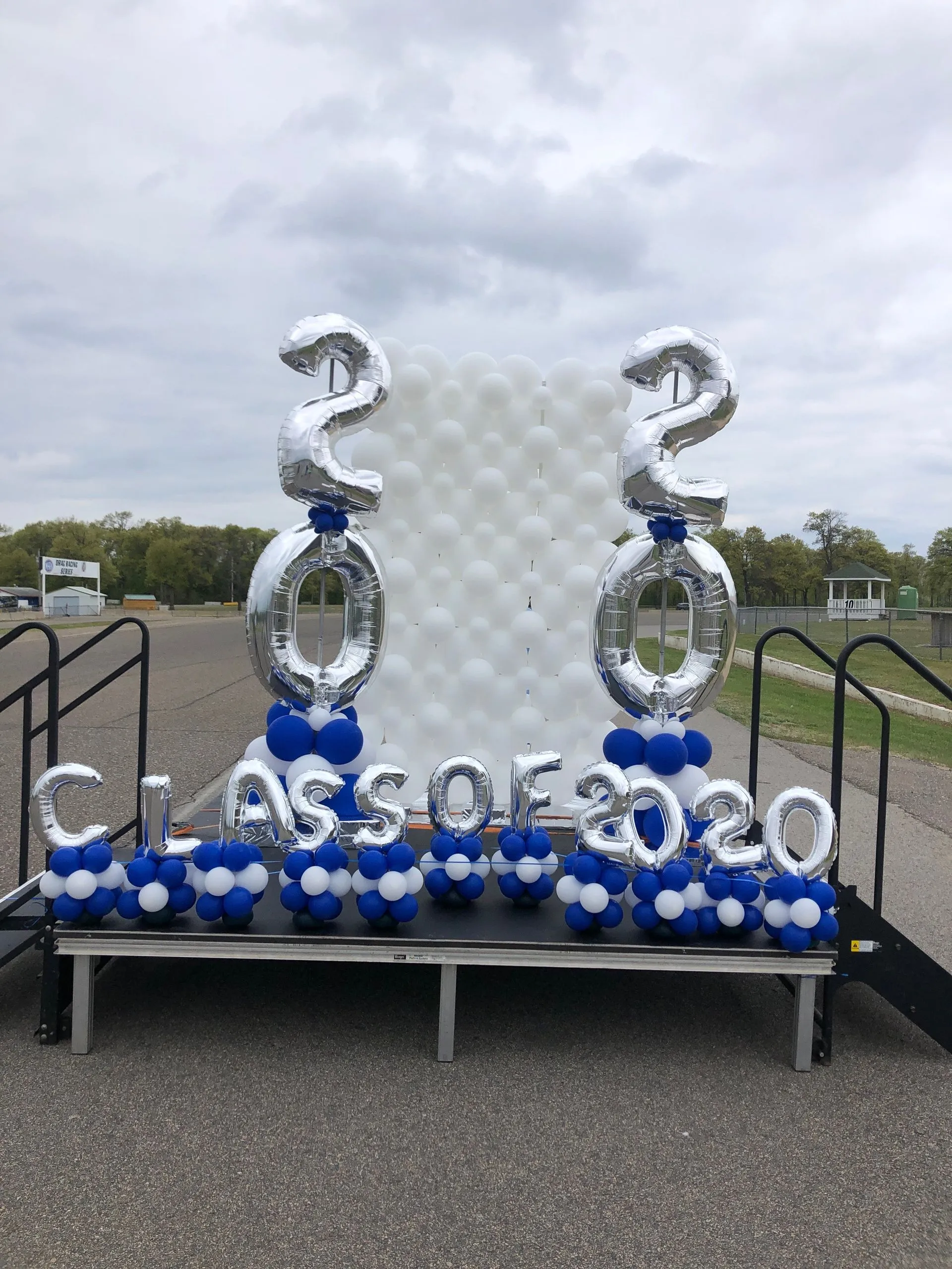 Outdoor graduation balloon display featuring blue and silver balloon columns with 2020 number toppers created by Be Bold Balloons