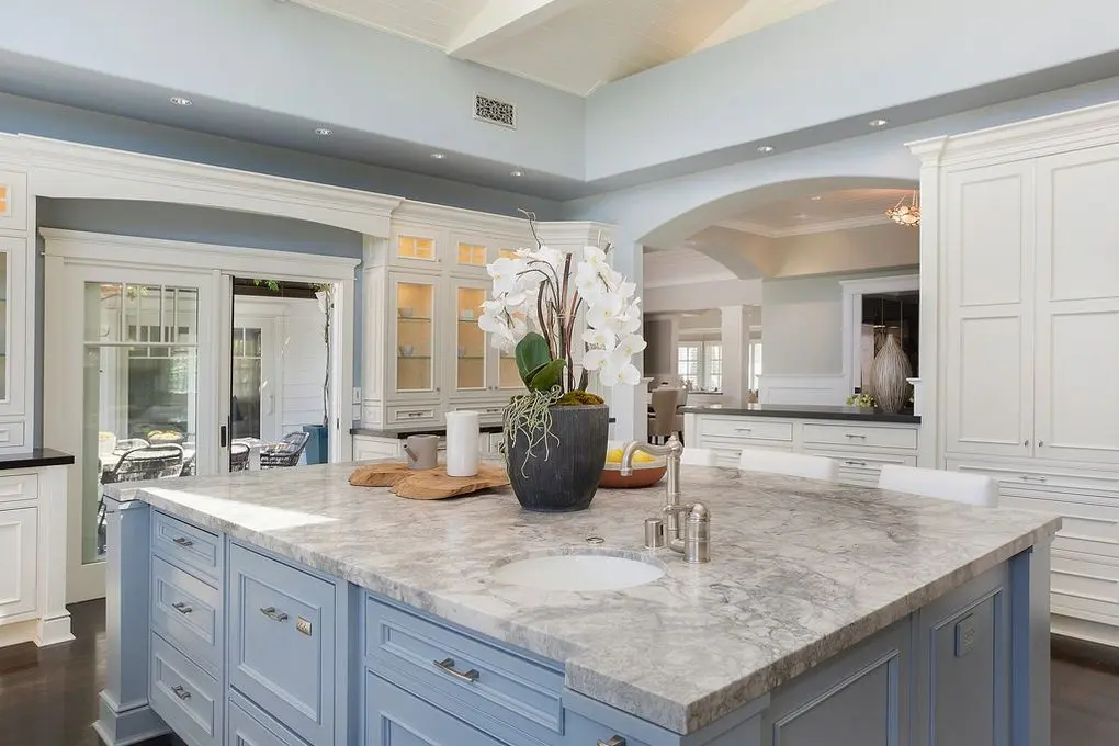 'Port Streets Remodel' kitchen with light blue island, marble top, white orchids, leading to a sunlit dining area through an archway.
