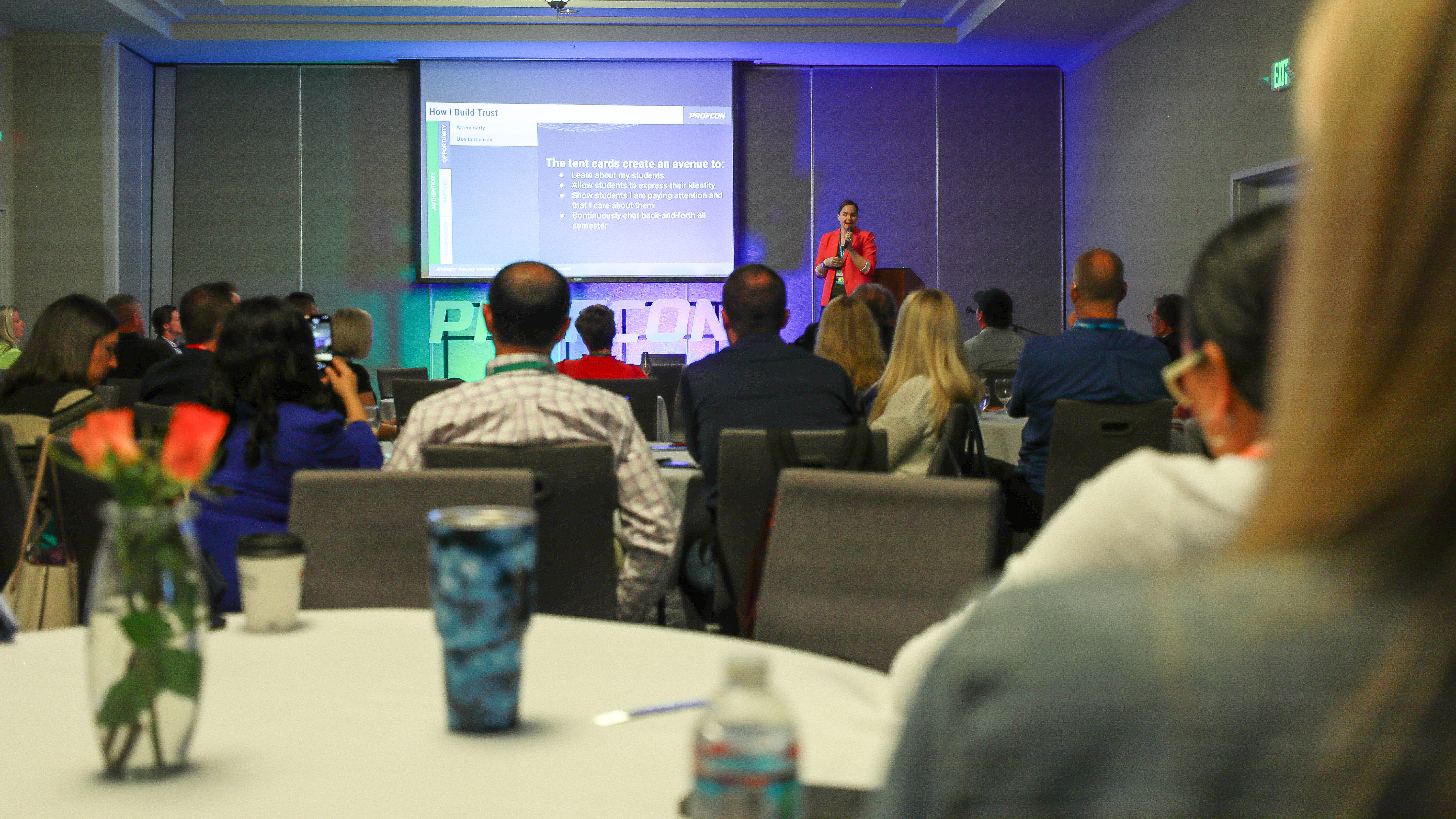 Speaker in red blazer presents at PROFCON conference to seated audience. Slide reads: “The best cards create an avenue to connect,” followed by a bulleted list. Tables with notebooks, water bottles, and flowers are visible in foreground.