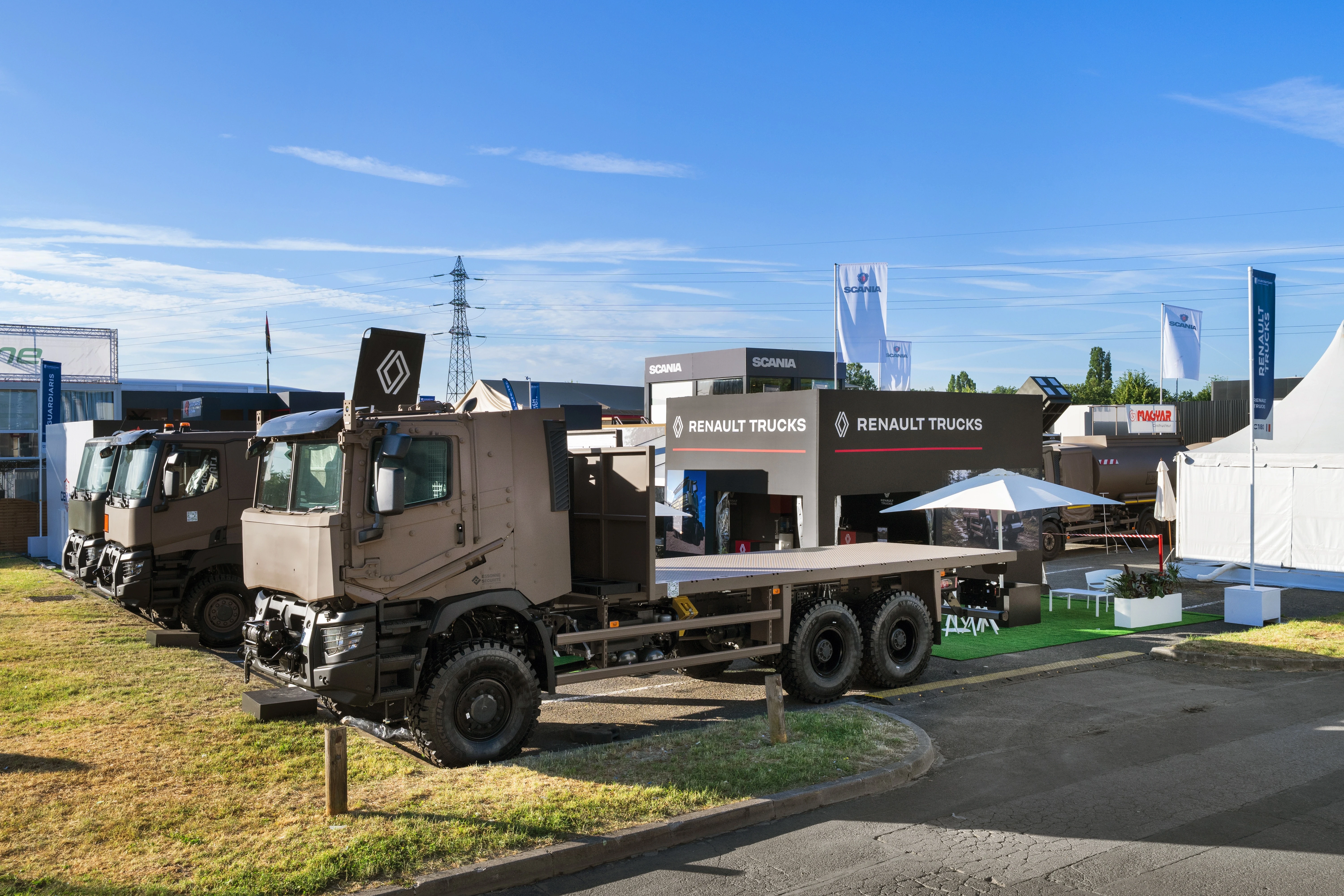 Stand extérieur Renault Trucks défense conçu par EXPACE à Eurosatory 2026 — camions militarisés 6x6 sable sur pelouse, façade anthracite et rouge, chapiteau d'accueil parquet