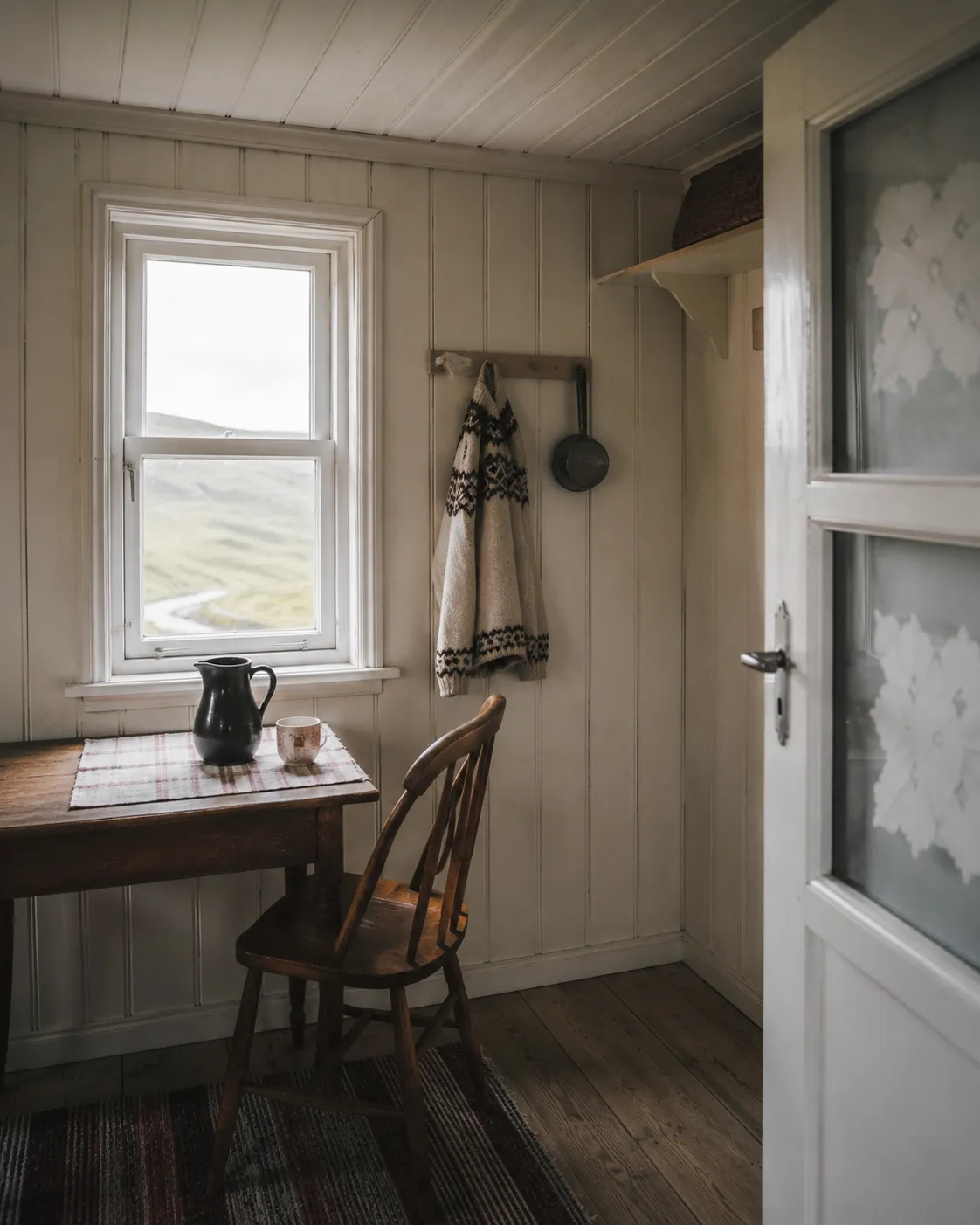 Cozy chalet dining corner with a wooden table, chair, white paneled walls, and countryside views through a window.