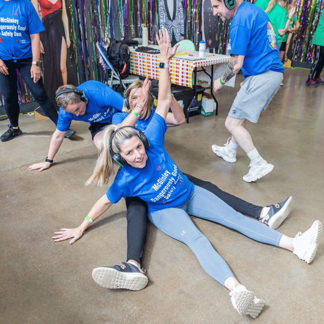 A group of people wearing blue T-shirts take part in an indoor activity. Two women are seated on the floor, laughing and raising their arms, while others around them join in the fun.