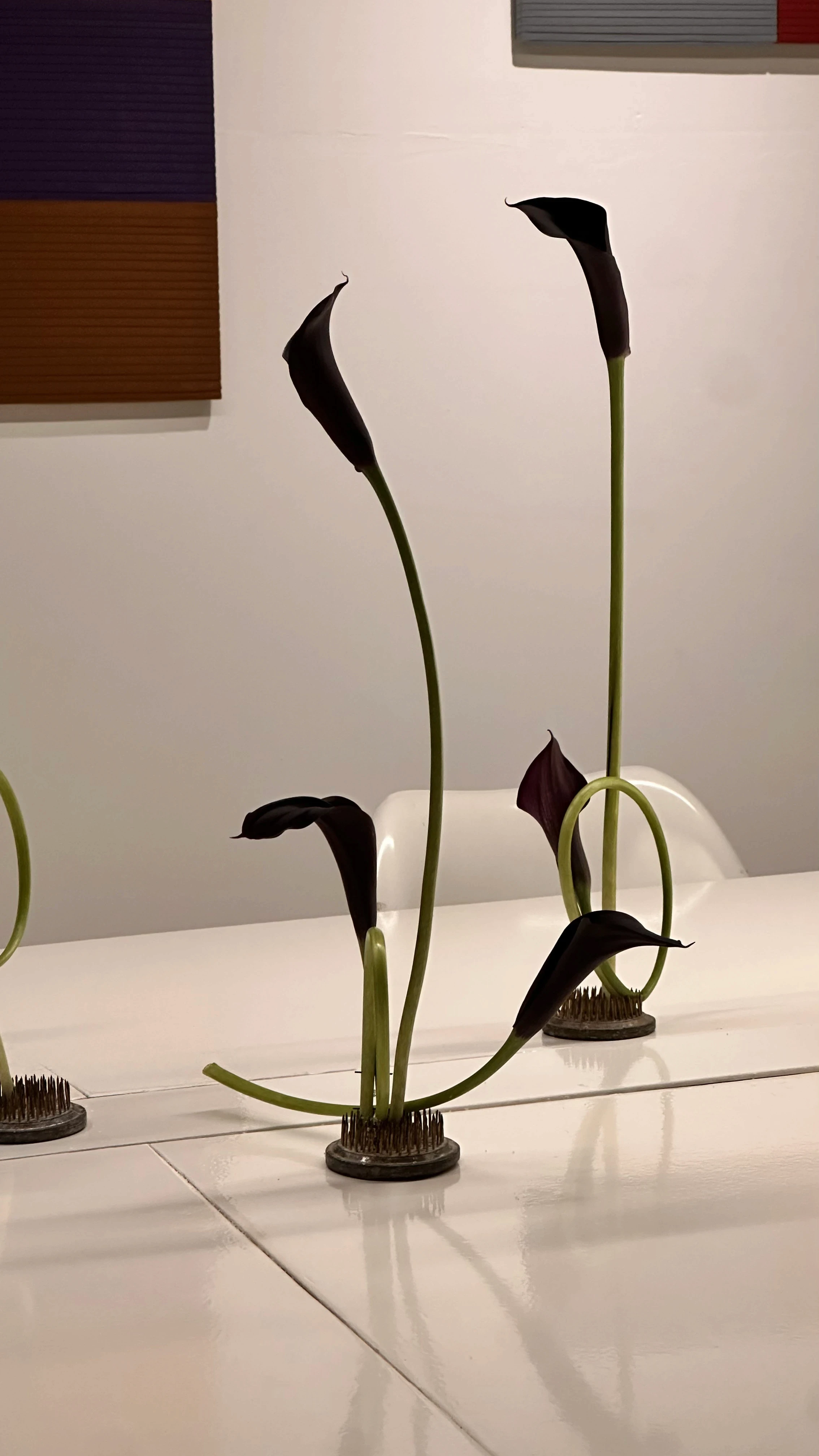 Calas negras de tallo largo en soporte kenzan, creando líneas minimalistas sobre una mesa blanca moderna.