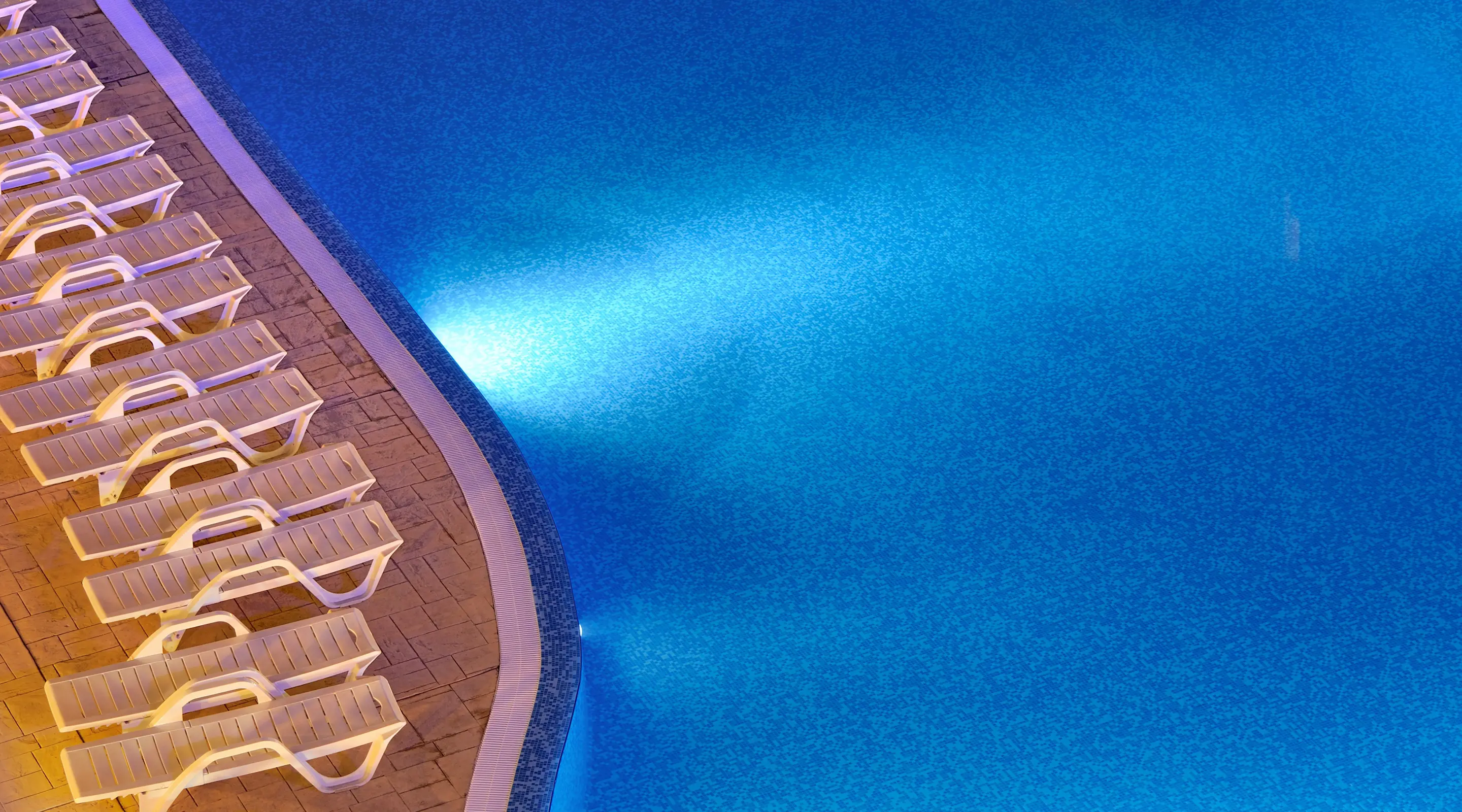 A clean pool at night with some resort chairs nearby